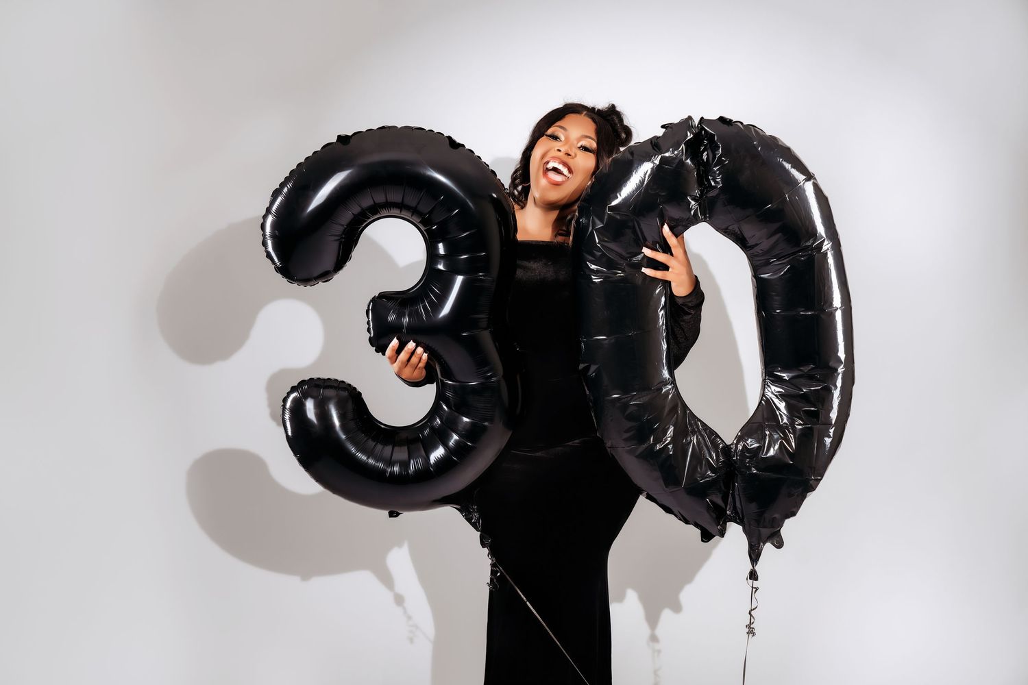 A woman in a black dress laughs while holding large black number 30 balloons against a white background.