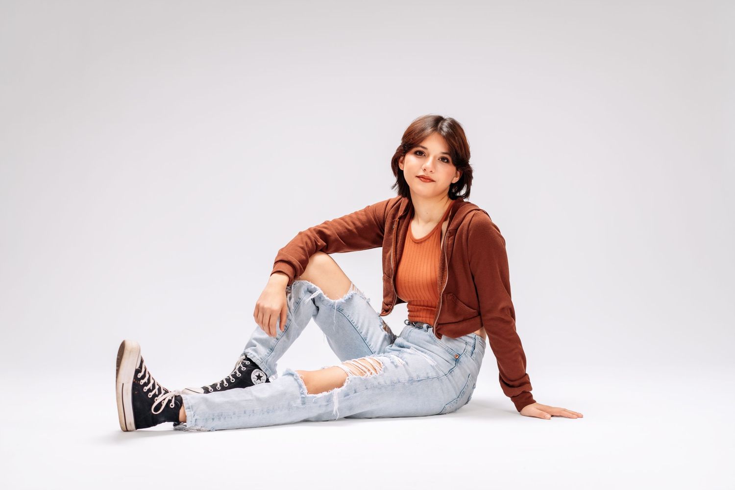 Young woman in ripped jeans, brown hoodie, and Converse sneakers sitting casually on a white studio floor.