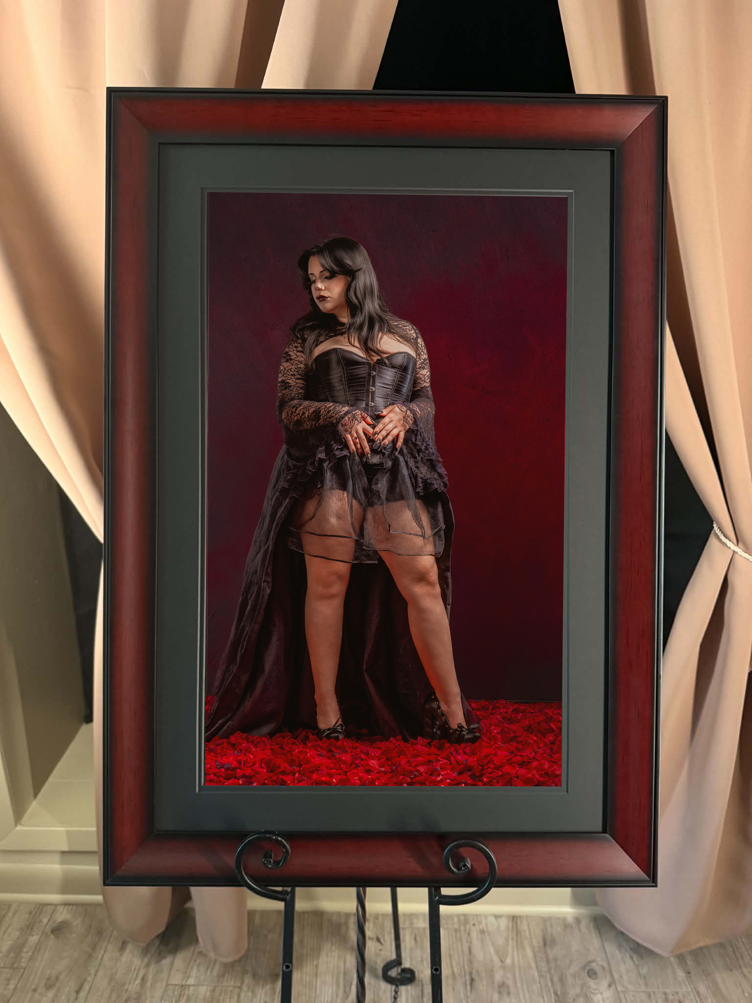 Framed fine art portrait of woman in dark lace ensemble standing on red roses on easel.