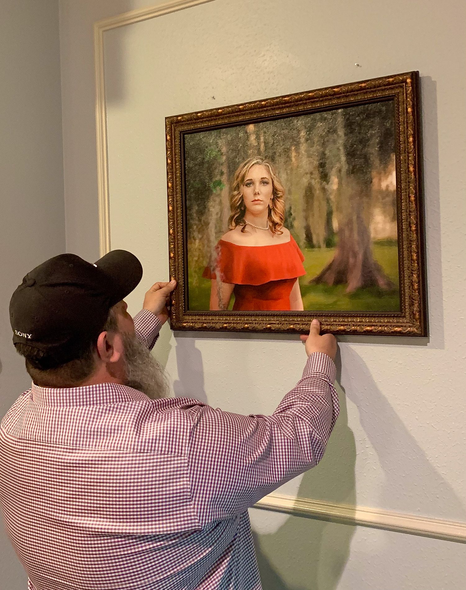 Man hanging a framed portrait painting of a woman in red dress on a white wall.