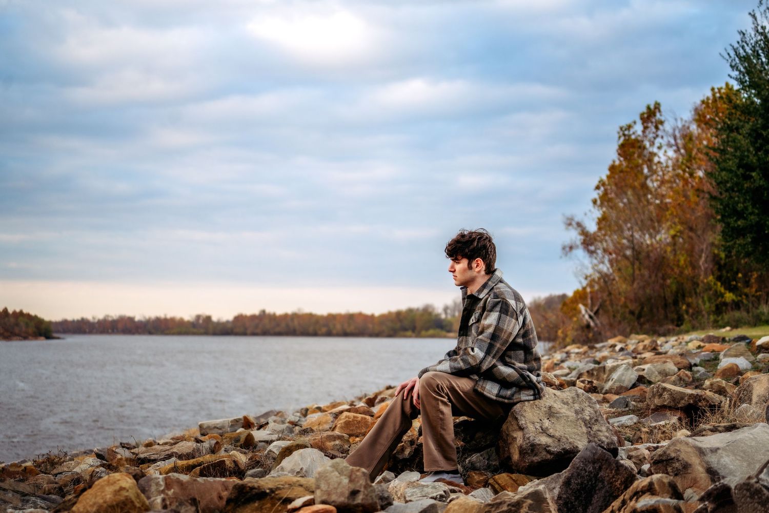 Young man sits contemplatively on rocky shoreline beside calm river with autumn trees.