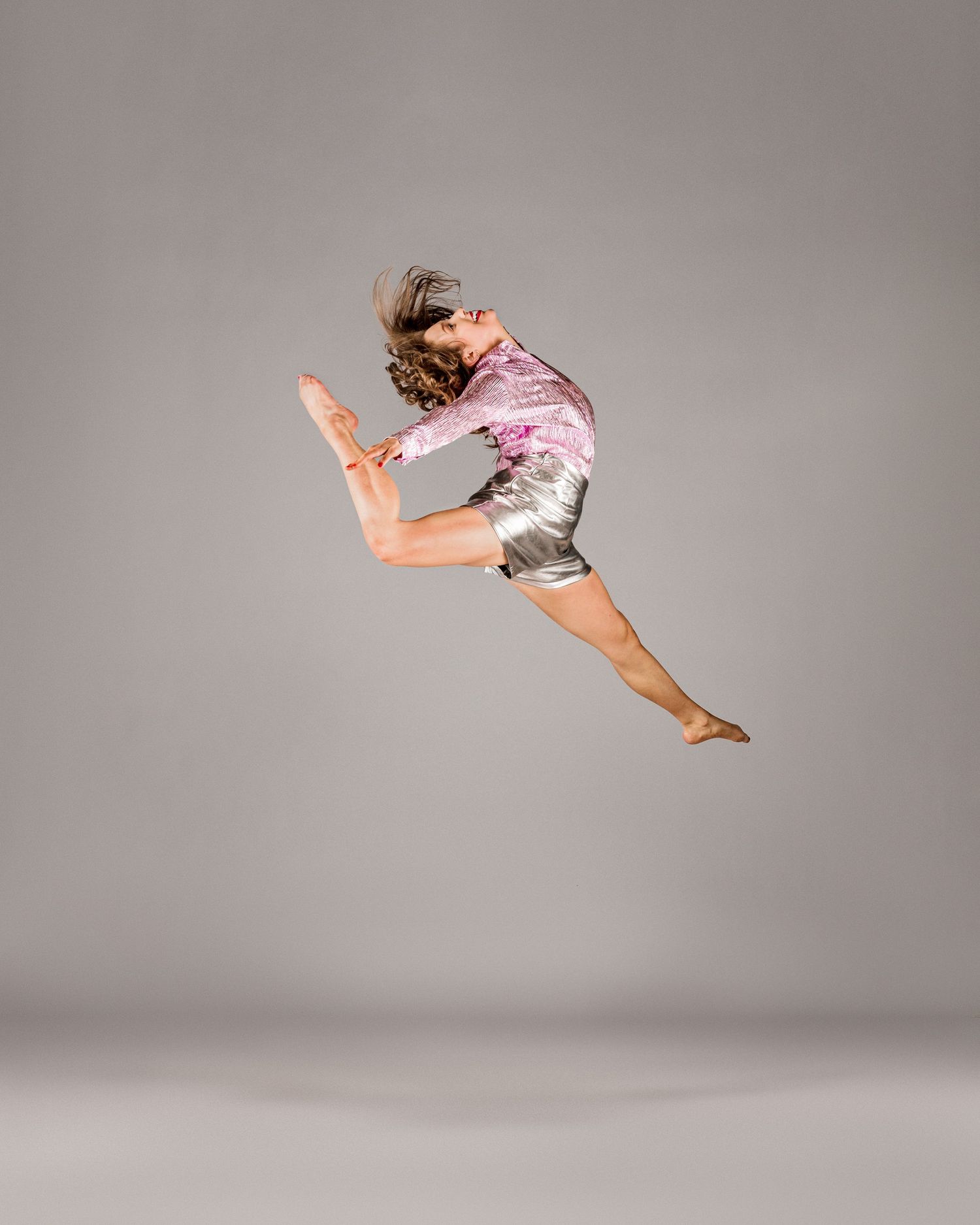A young female dancer leaps gracefully in a pink and silver costume against a gray studio background.