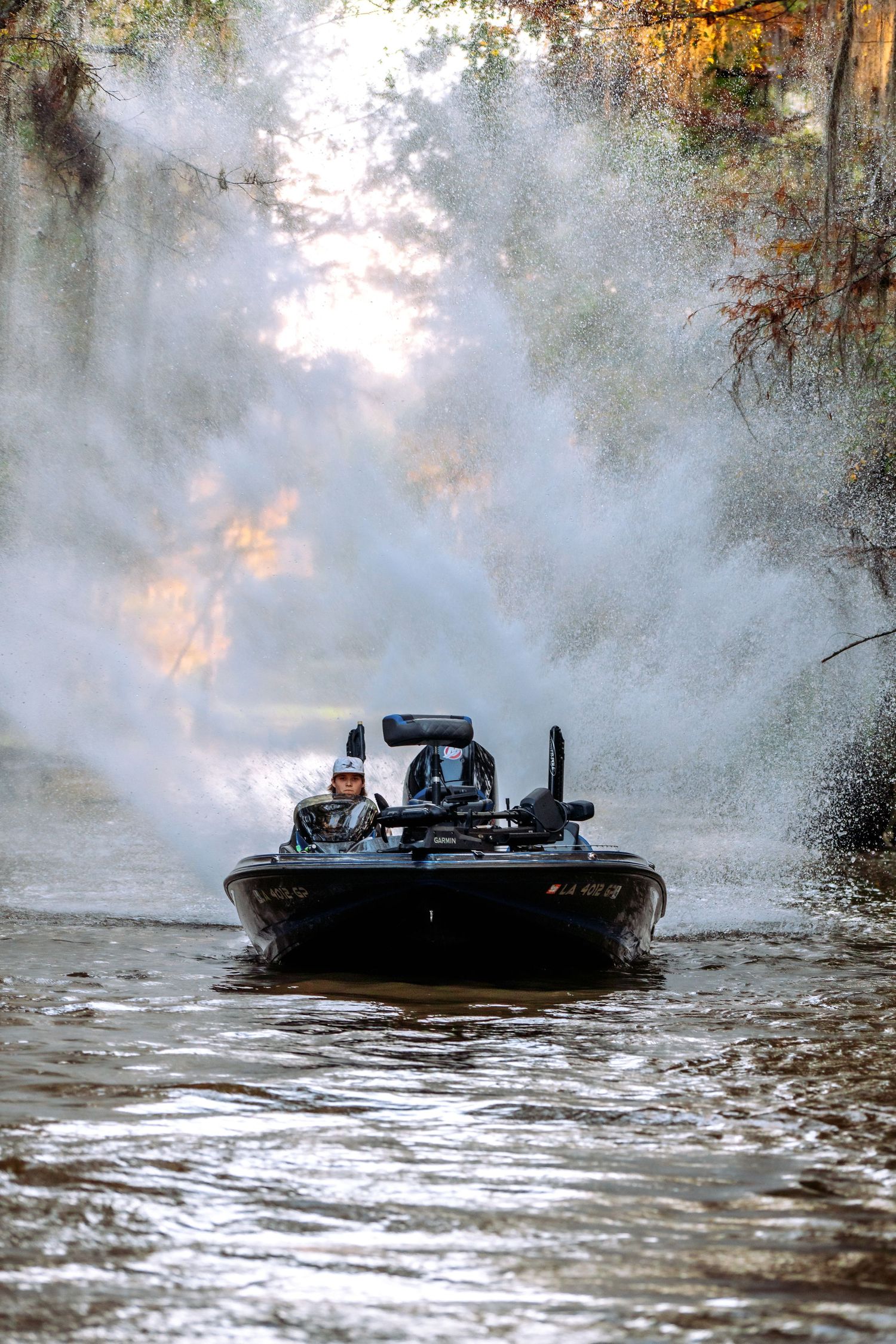 A bass fishing boat speeds through a misty river surrounded by trees, creating dramatic water spray in golden morning light.