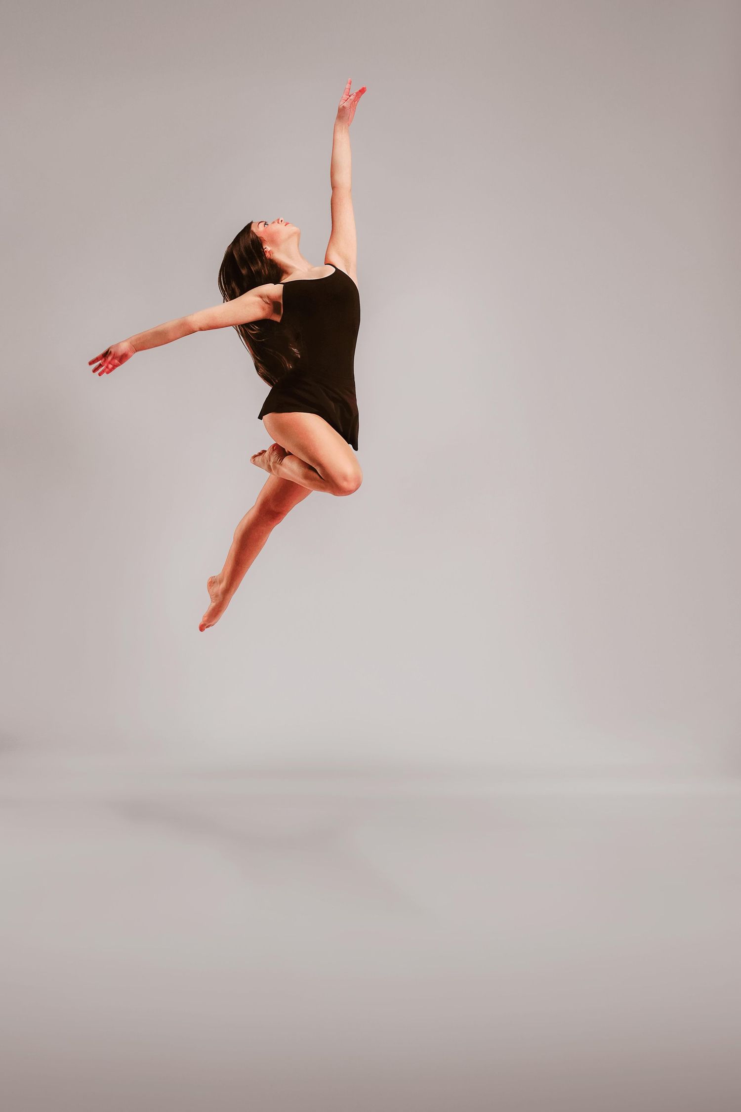 A female dancer leaps gracefully in a black leotard against a gray studio background, arms and legs extended mid-air.