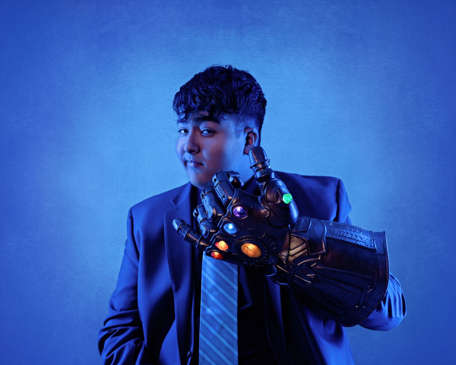 Young man in blue suit wearing Infinity Gauntlet prop under dramatic blue studio lighting.