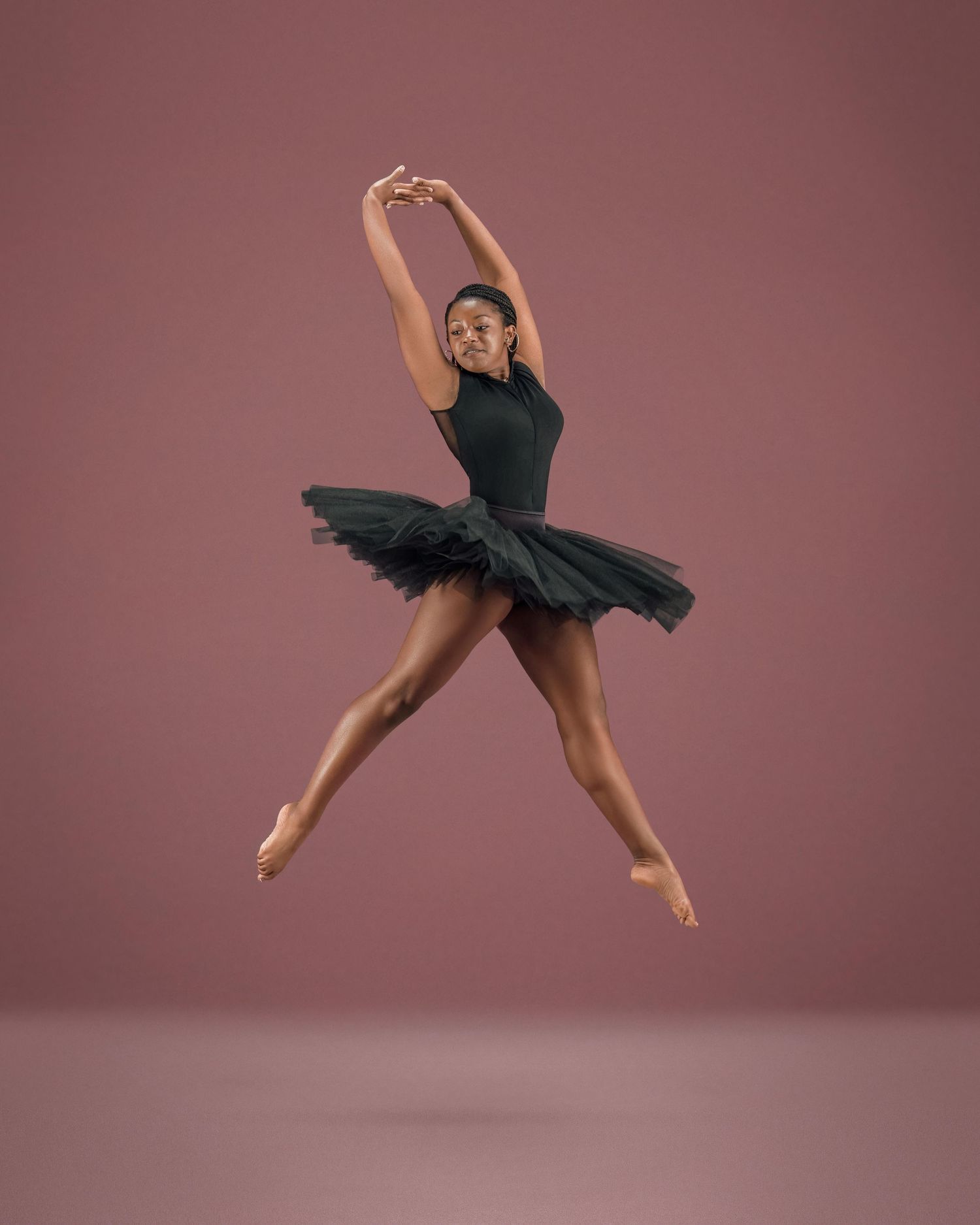 A ballet dancer in a black tutu leaps gracefully against a mauve background, arms raised overhead.