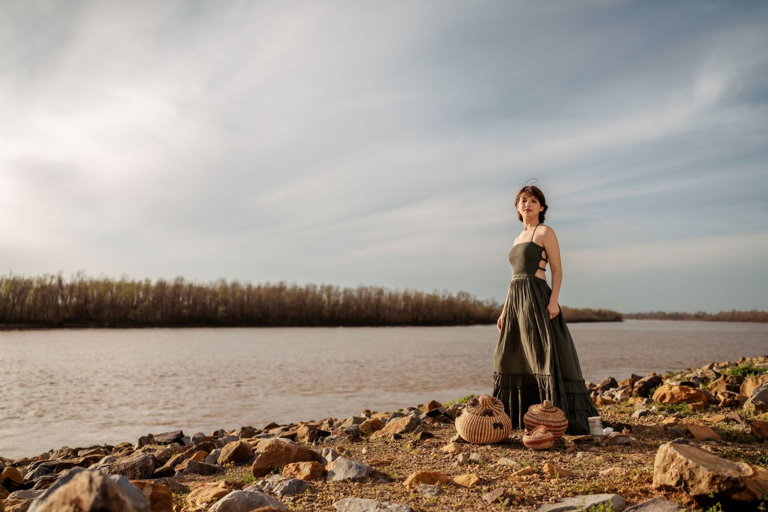 Woman in olive green dress standing on rocky shore by a river with trees in background under cloudy sky.