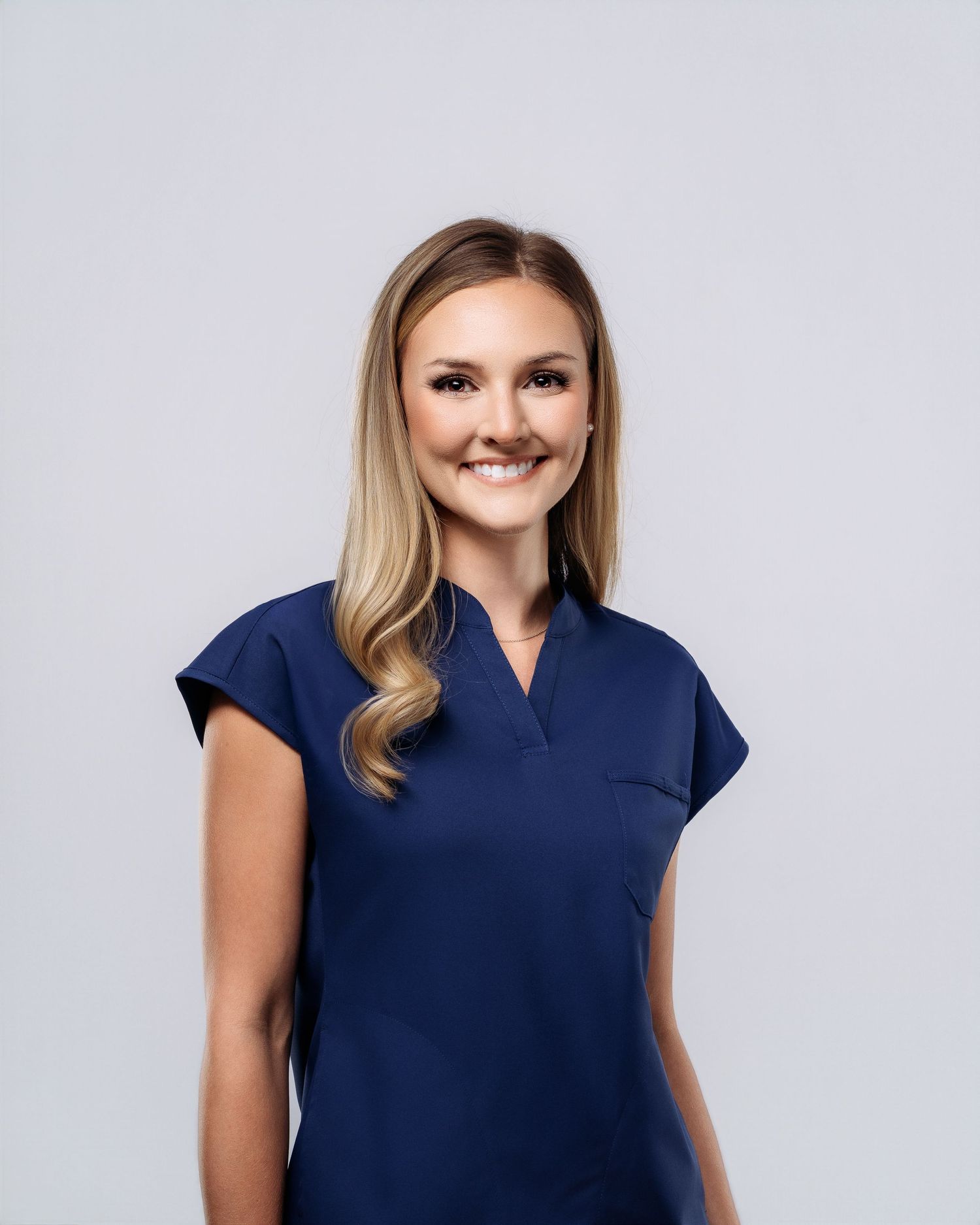 Smiling woman with blonde hair wearing a navy blue scrub top, posing against a light gray background.