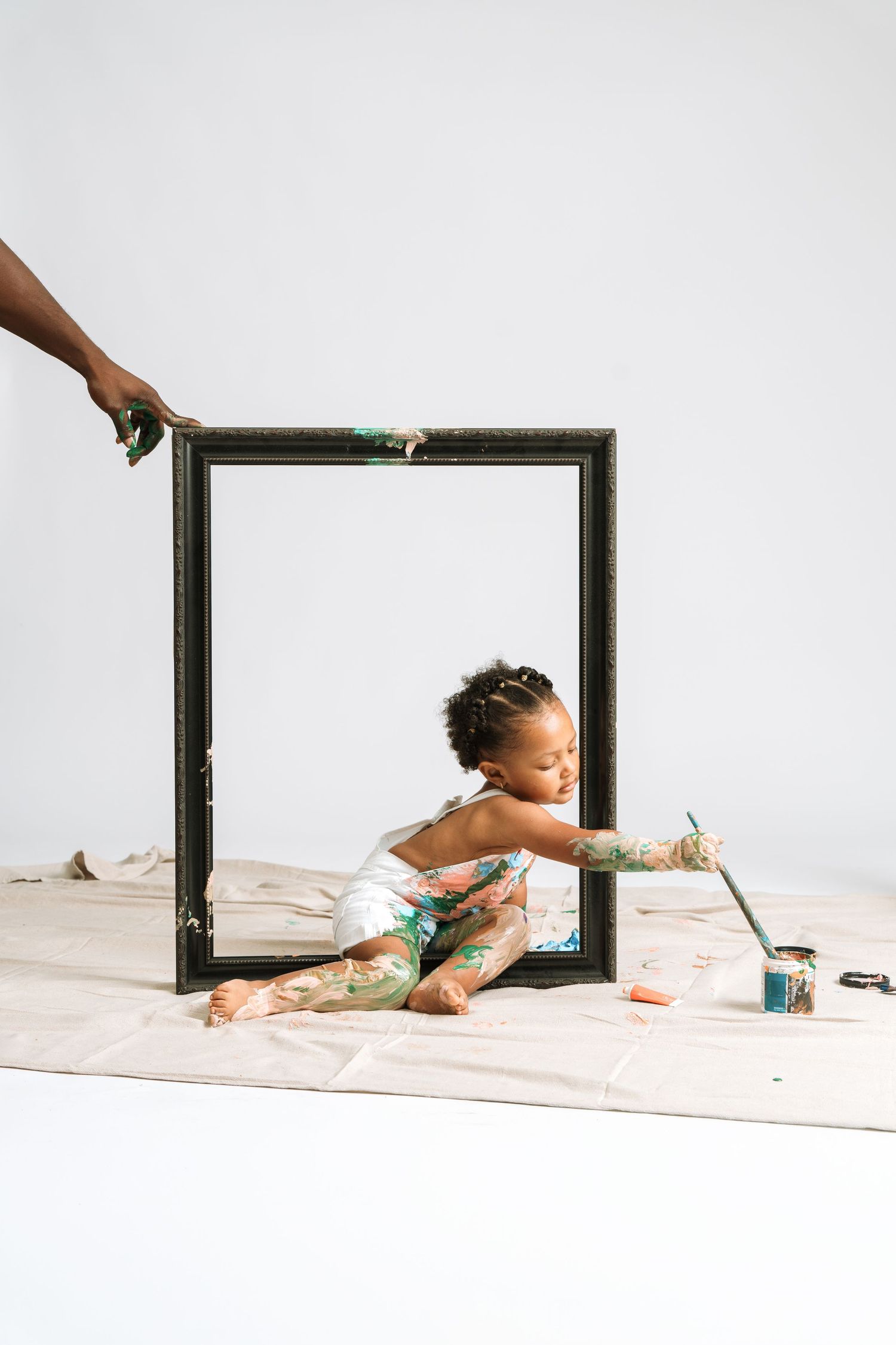 A toddler covered in green paint sits inside a large black frame, holding a paintbrush, on a drop cloth.