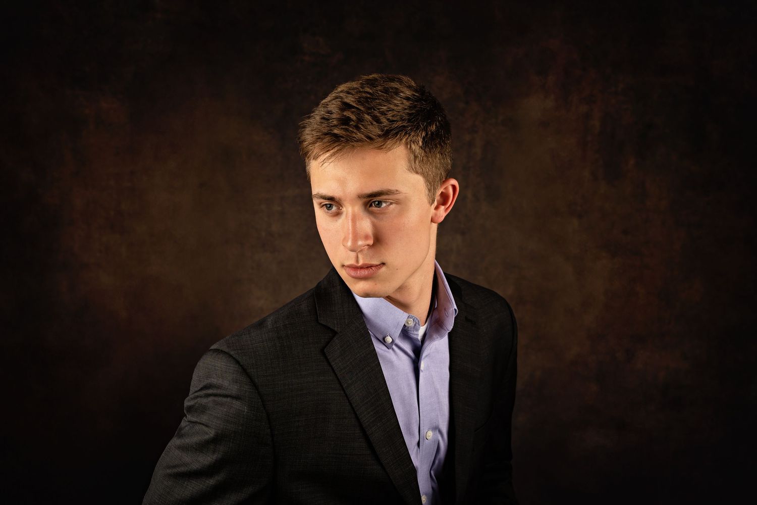 Young man in dark blazer and blue shirt poses against a dark brown studio background for a professional portrait.