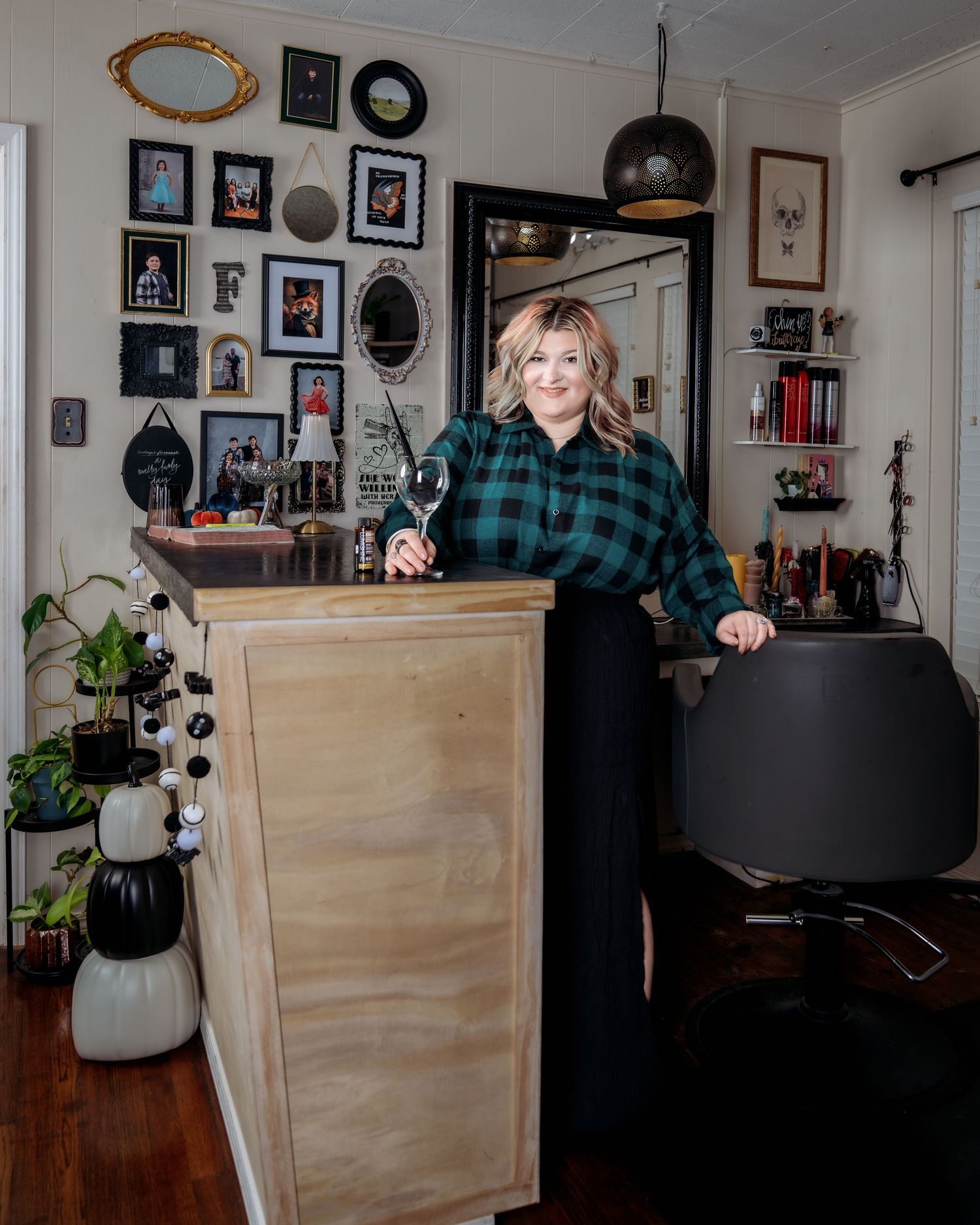 Woman in plaid shirt holding wine glass stands beside wooden reception desk in cozy salon with gallery wall.