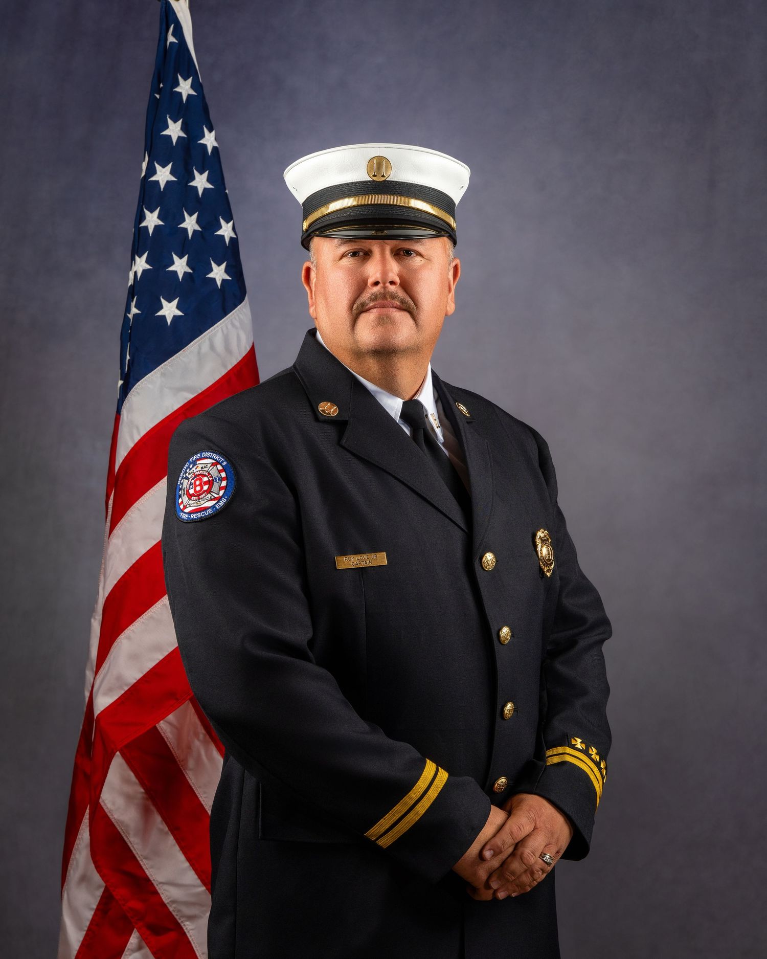 Fire department officer in dress uniform with gold insignia poses beside an American flag for an official portrait.