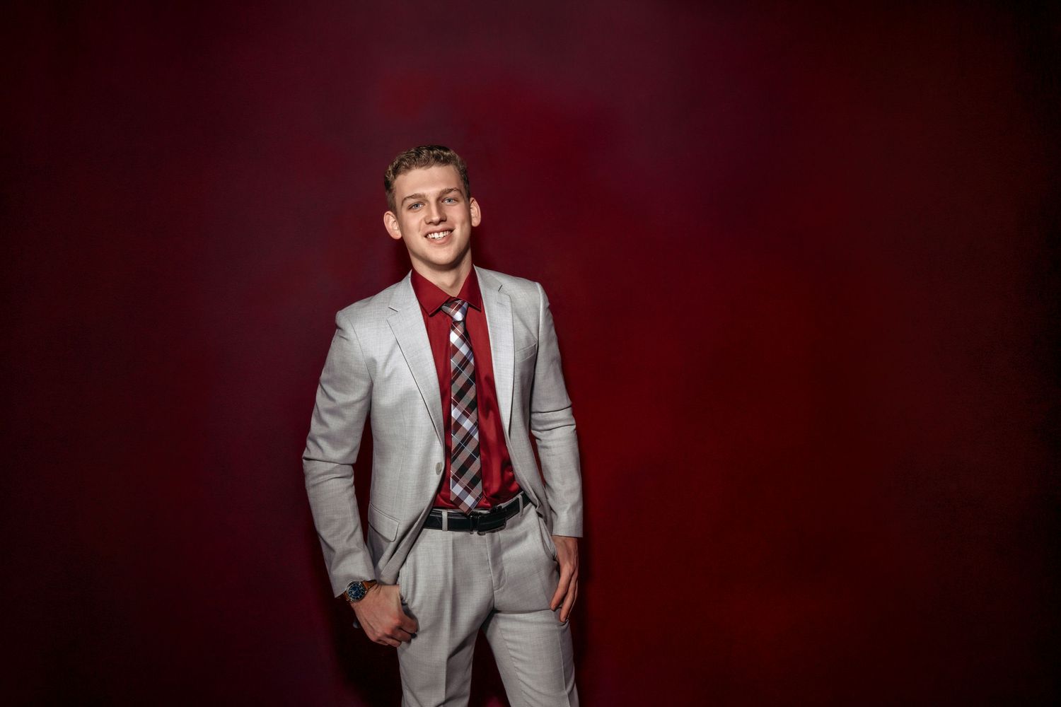 Young man smiling in a light gray suit with red shirt and striped tie against a dark red background.