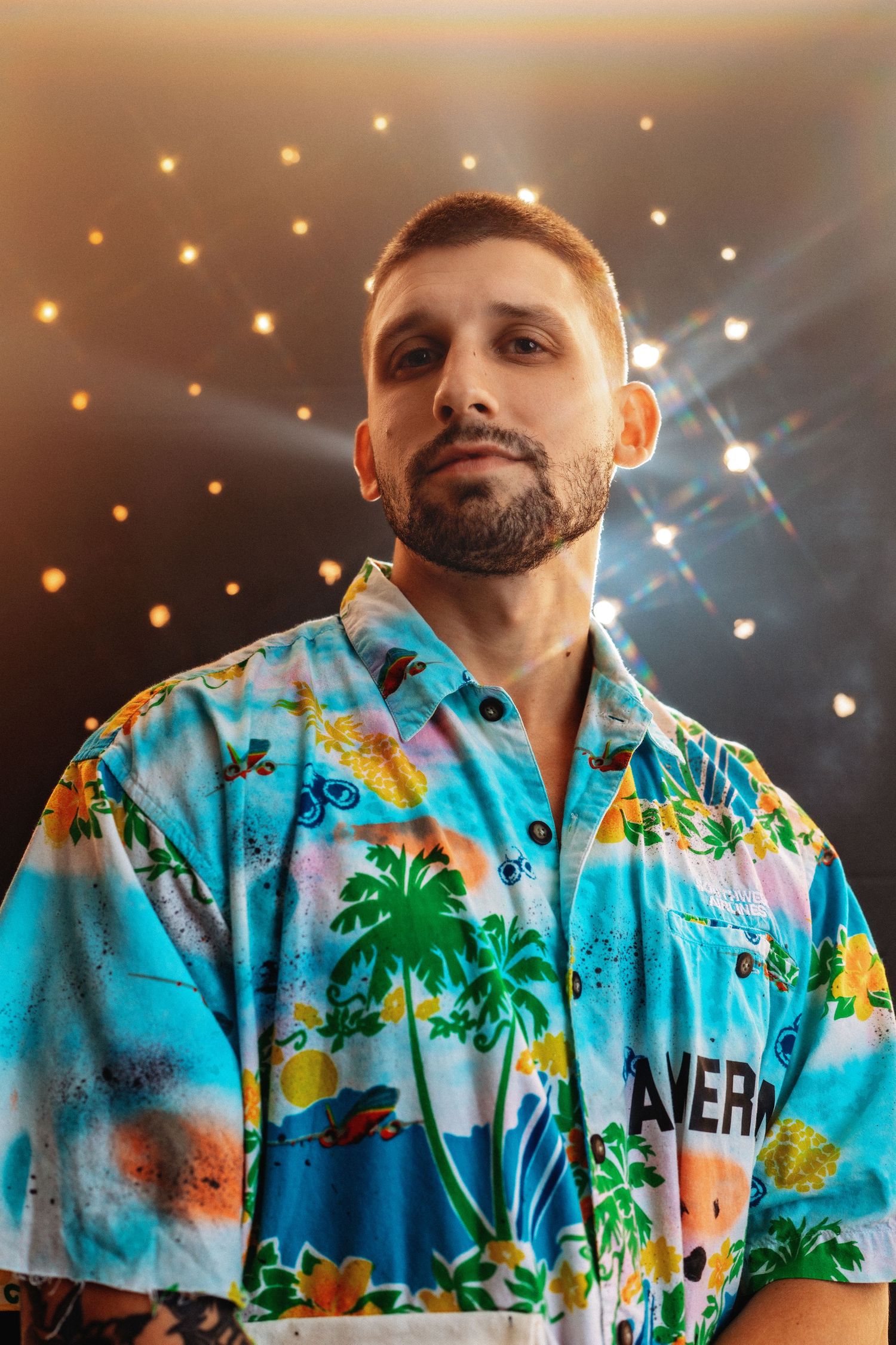 Bearded man in colorful tropical Hawaiian shirt posing under stage lights.