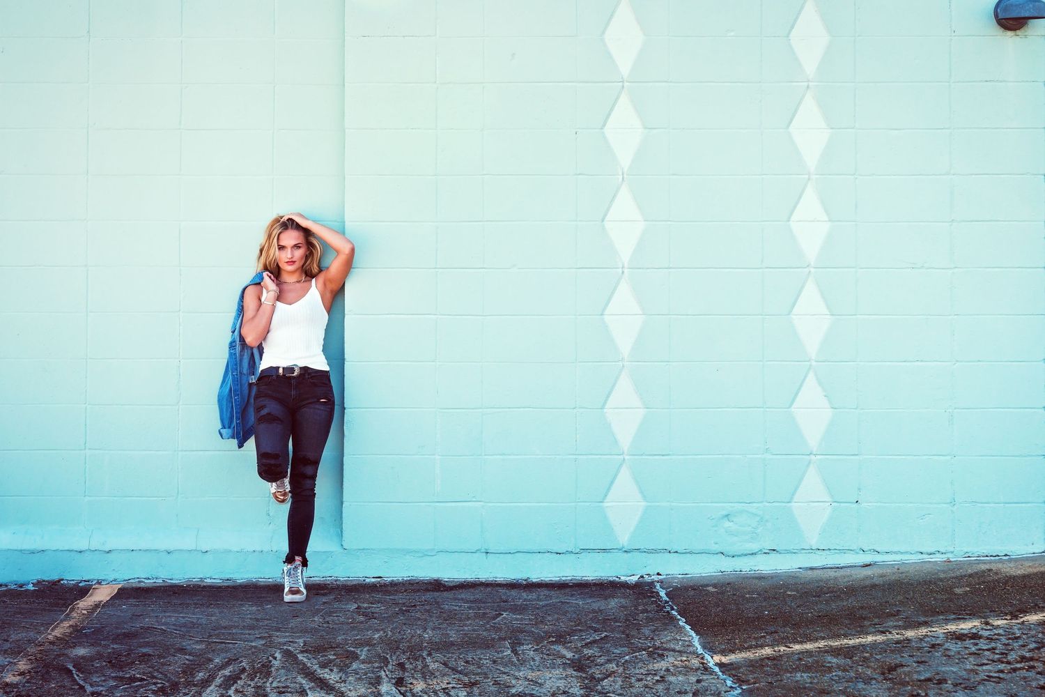 Young woman in white top and black jeans leans against a light blue wall wearing a sun hat.