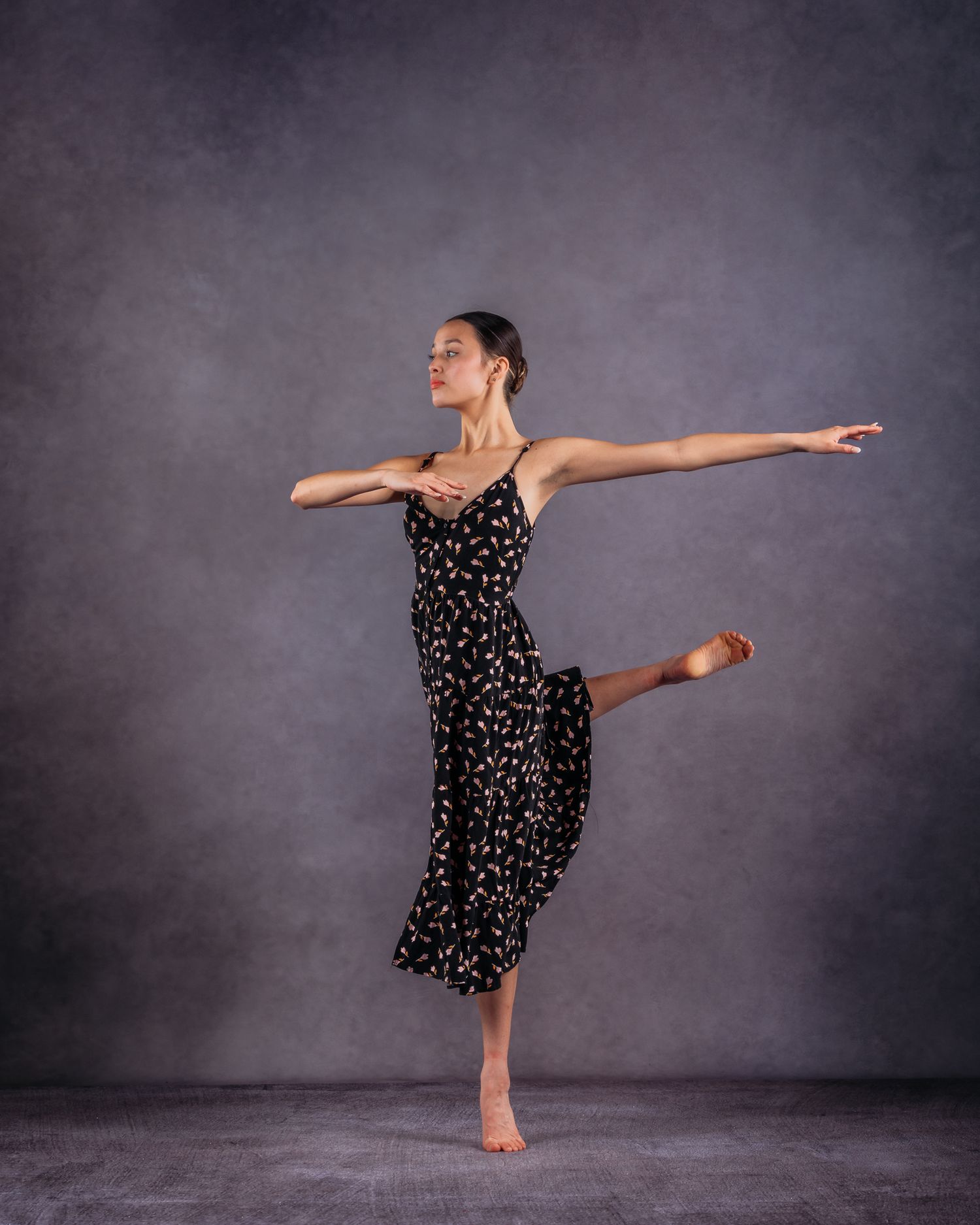 Female ballet dancer in a black floral dress balancing on one leg with arms extended gracefully.