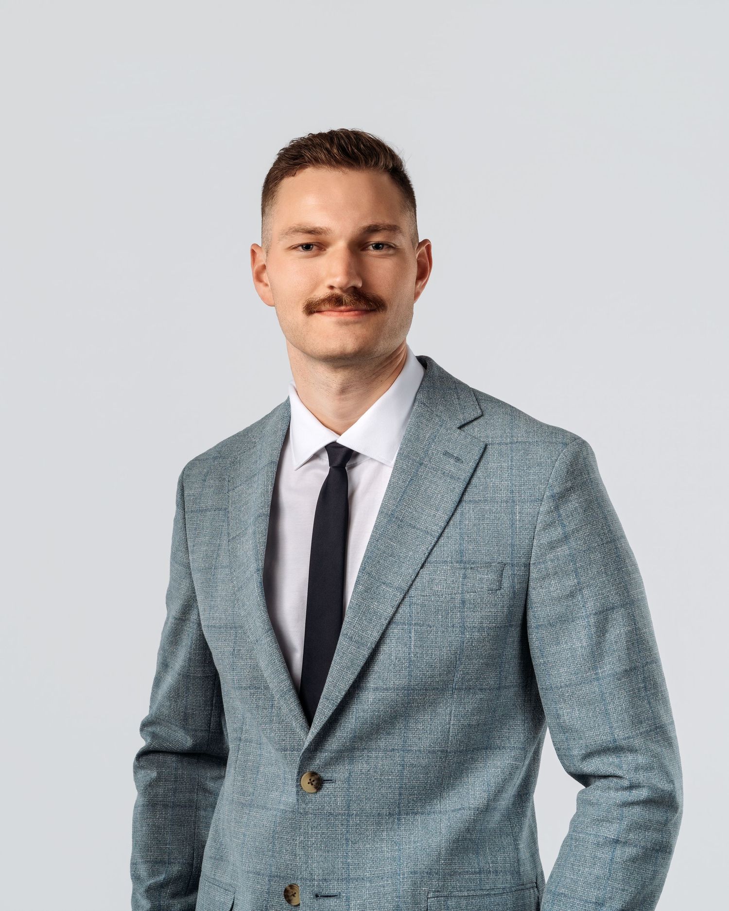Professional man with mustache wearing a light blue plaid suit and dark tie poses for a corporate headshot.