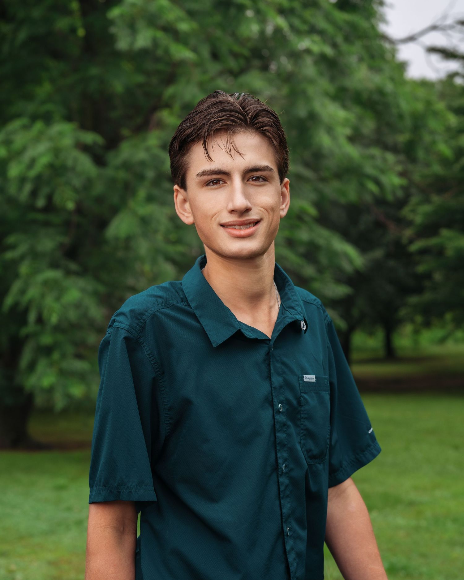 Young man in teal button-up shirt smiling outdoors with lush green trees in background.