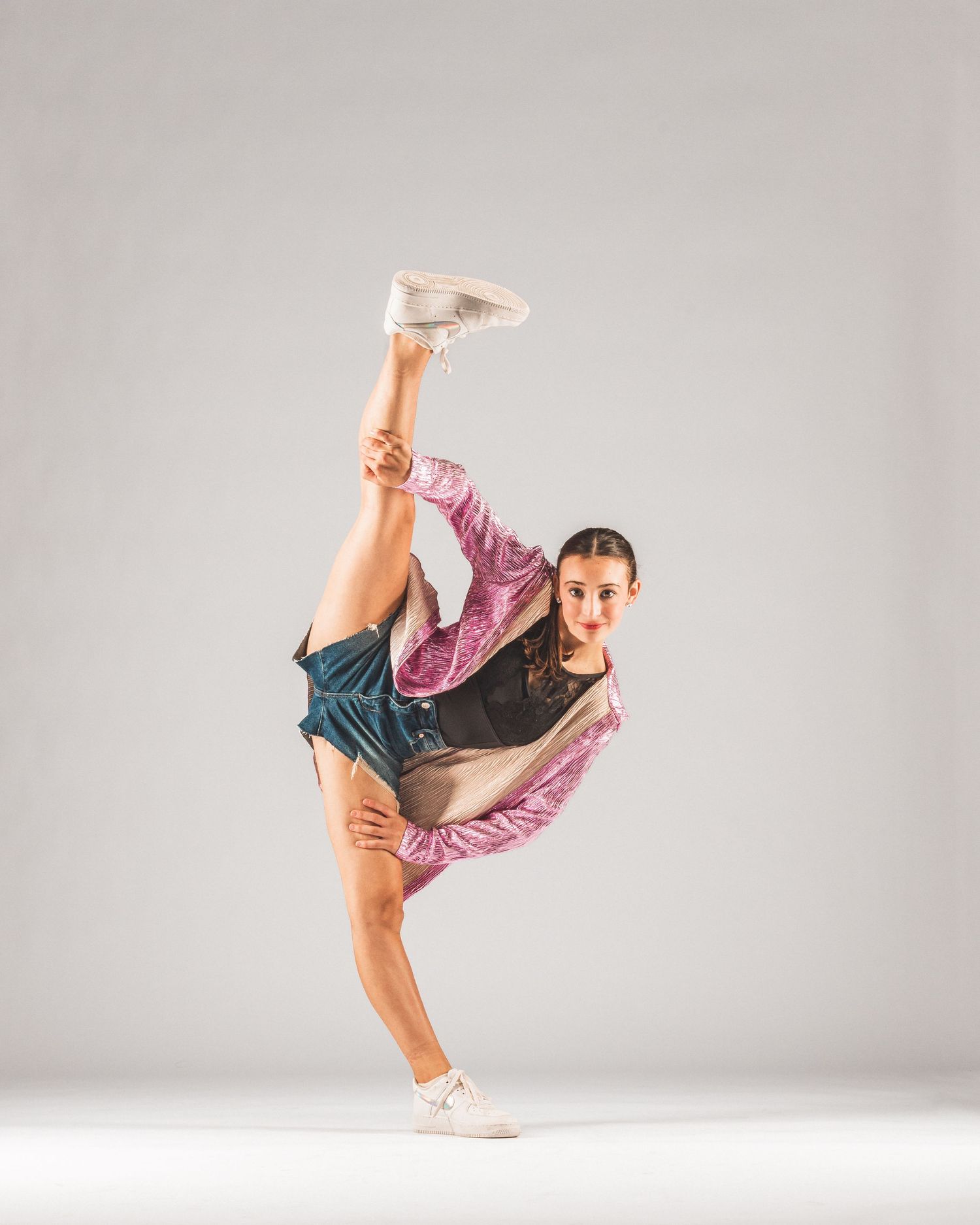 A flexible young dancer in a pink jacket and denim shorts performs a high kick in a studio.