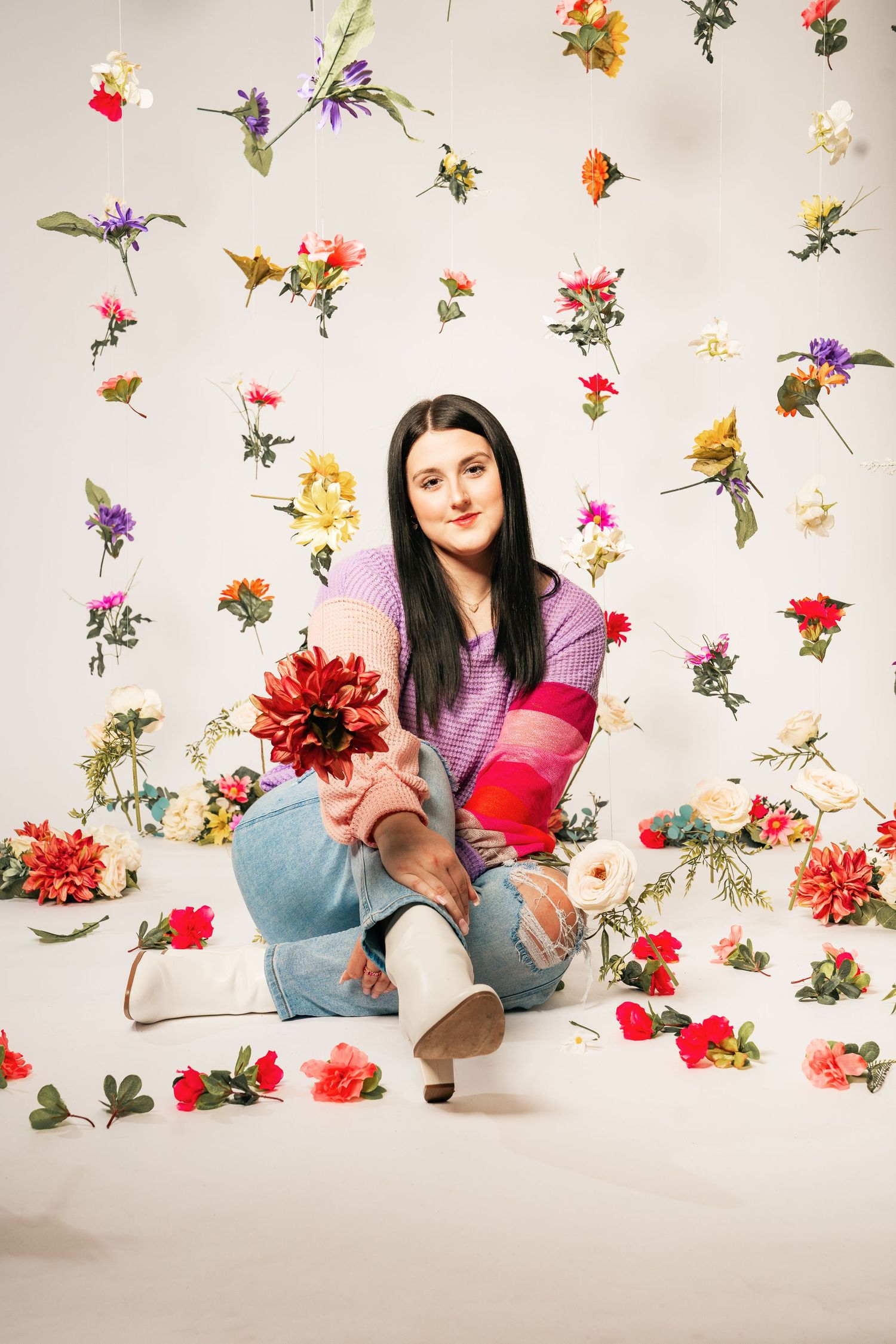 Woman sitting on floor holding red dahlia, surrounded by colorful hanging and scattered flowers on white background.