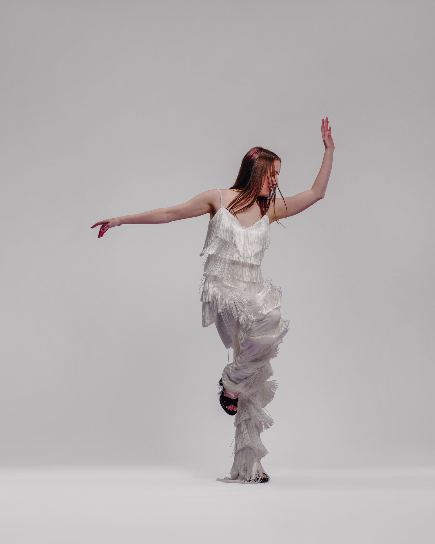 A woman in white ruffled outfit poses gracefully mid-dance against a light gray studio background.