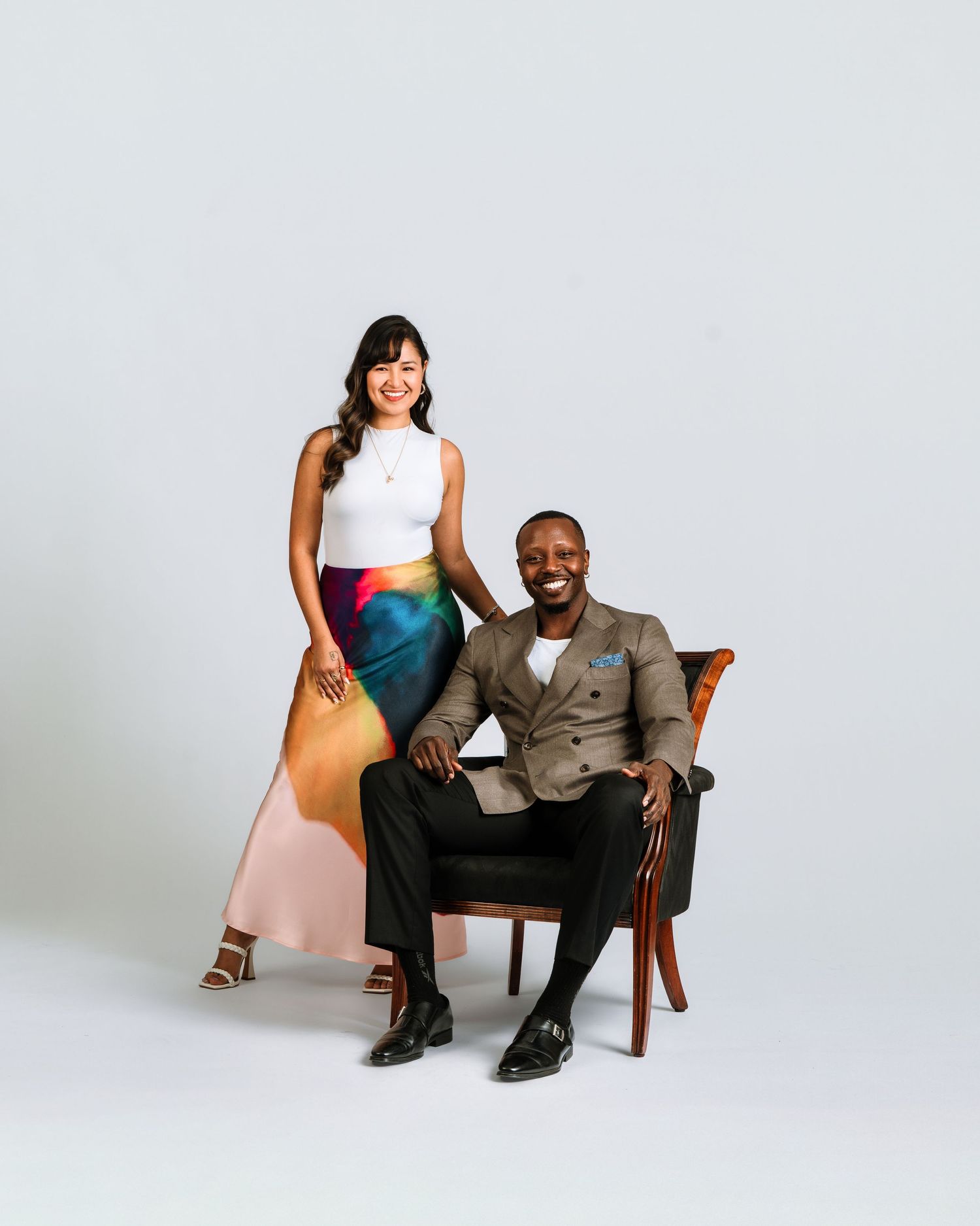 A smiling man in a suit sits in a chair beside a woman in a white top and colorful skirt during a professional photo shoot.