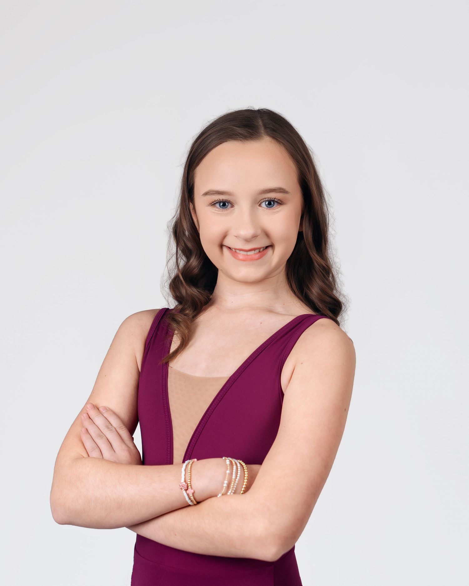 Young girl in burgundy sleeveless dress with arms crossed, smiling confidently against a light gray background.