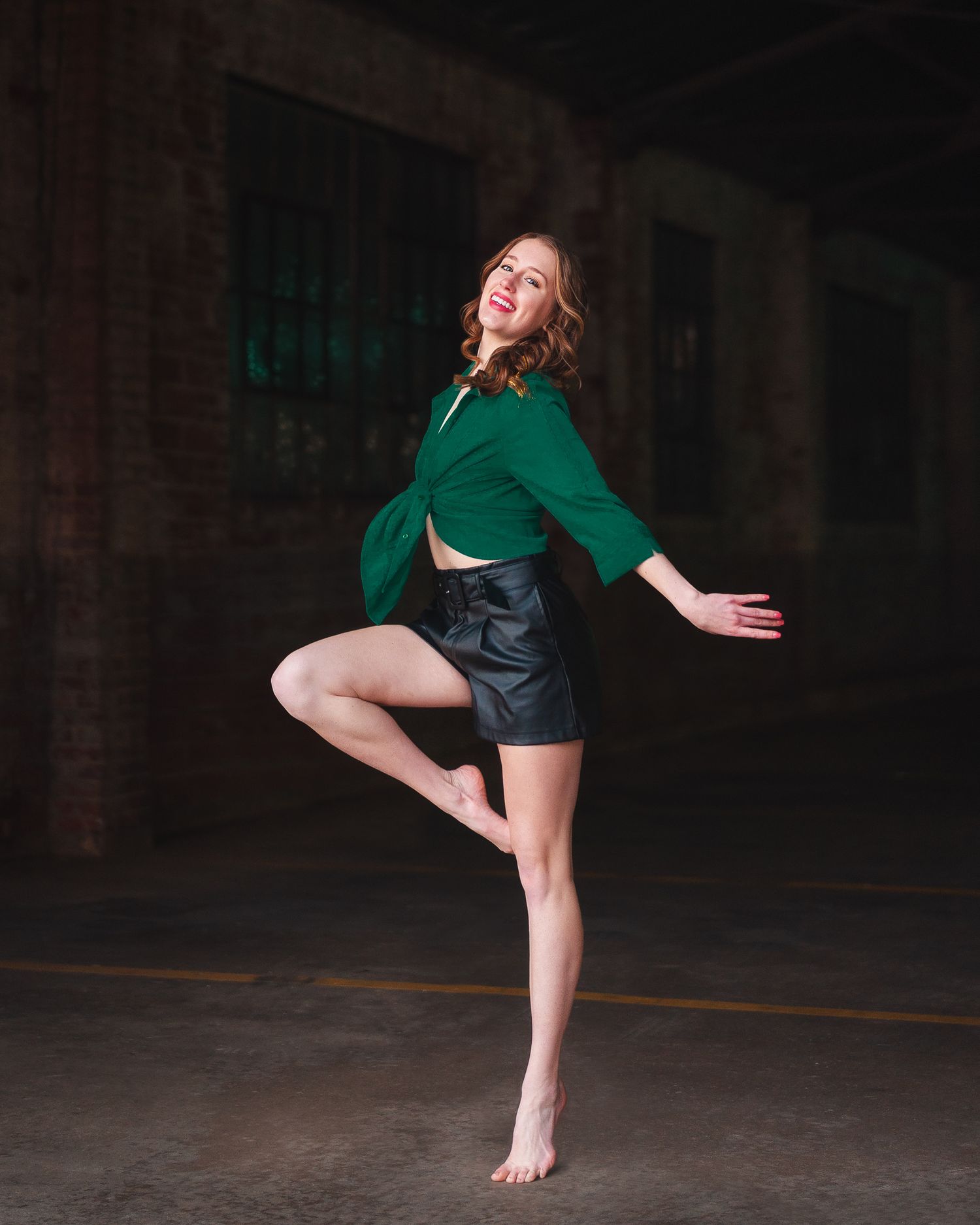 Female dancer in green top and black shorts leaps gracefully in dramatic industrial warehouse.