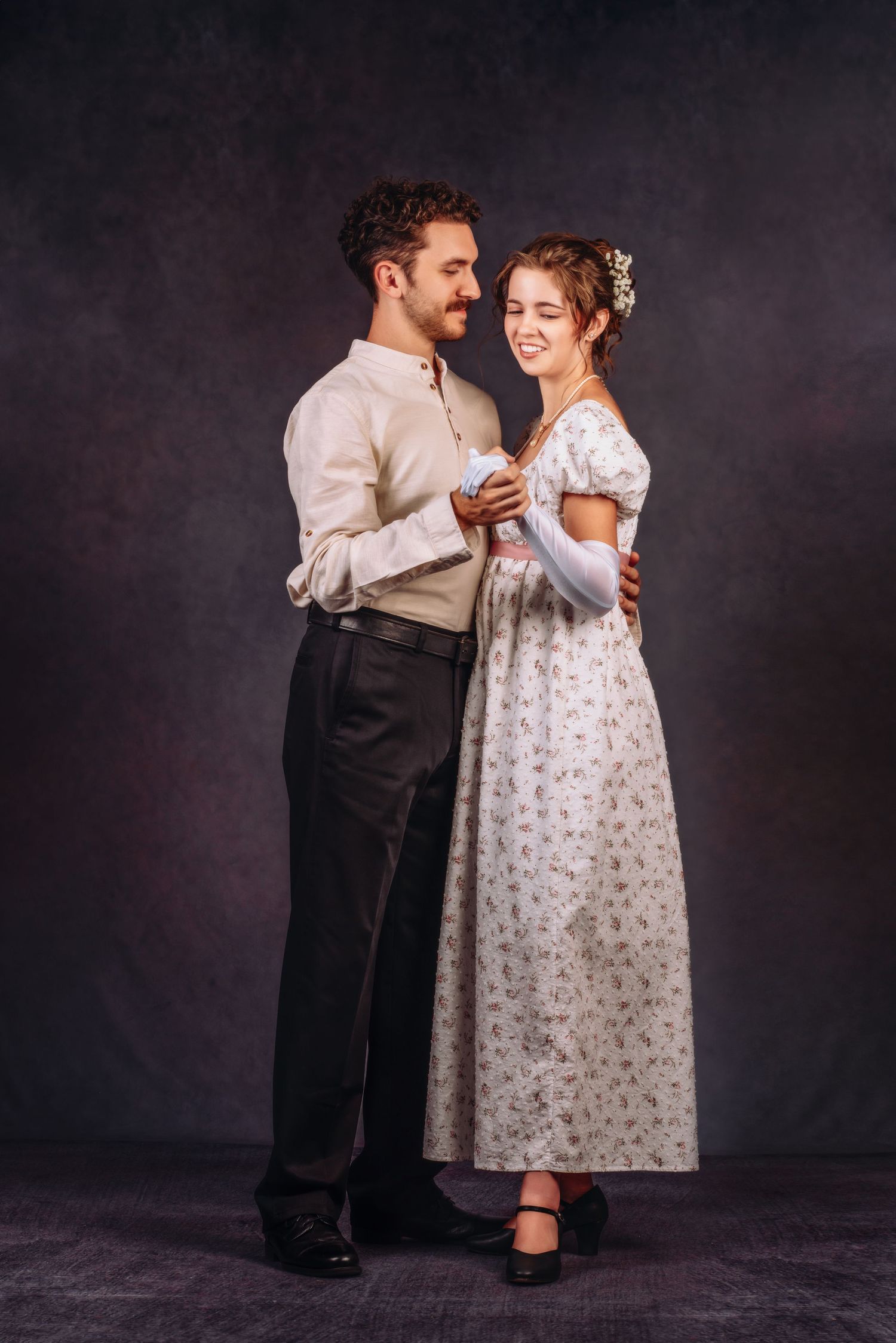 A couple in period costumes dance together, the woman in a floral Regency dress with flowers in her hair.