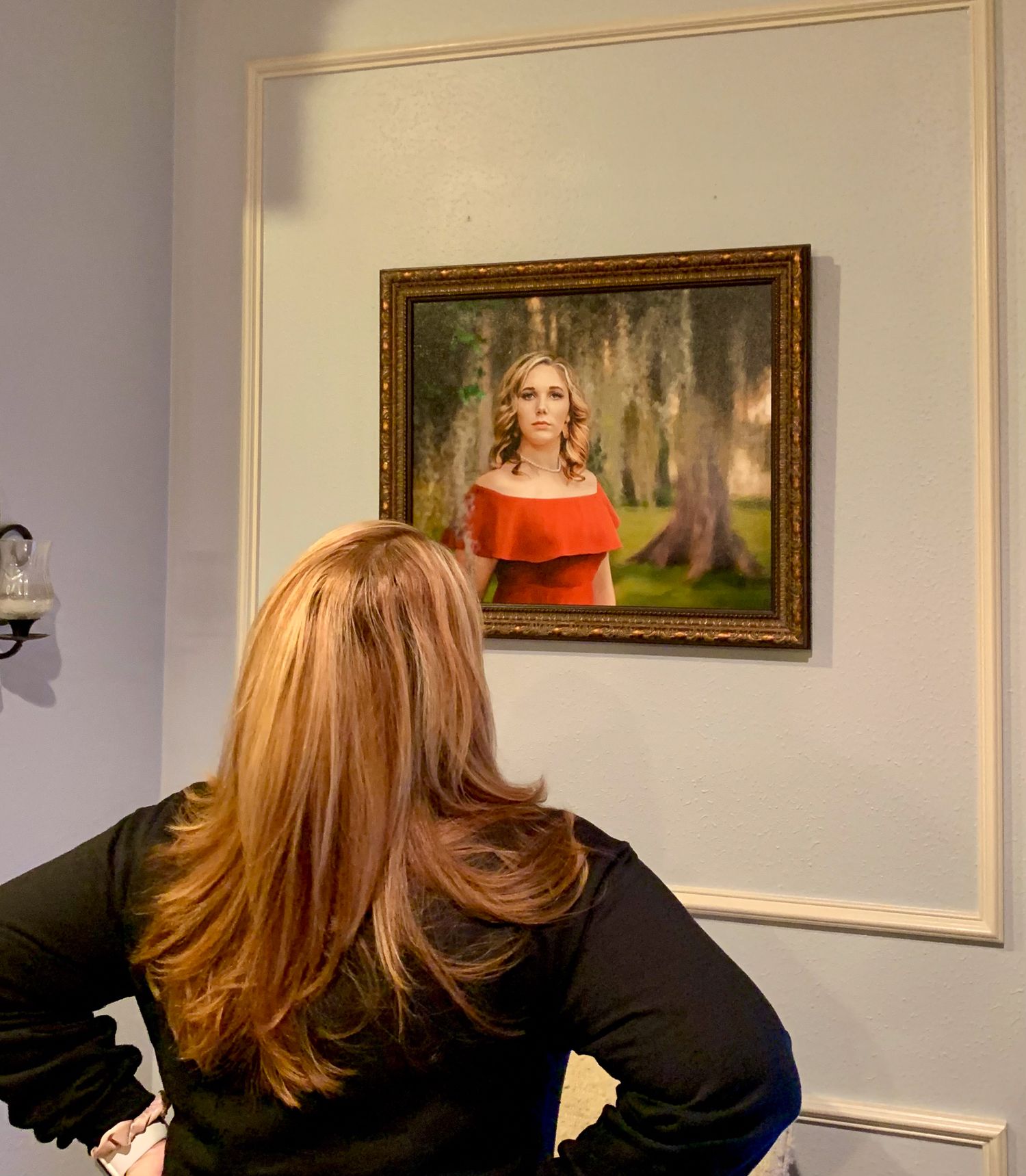 Woman with blonde hair viewing a framed portrait painting of a woman in red dress on a gallery wall.