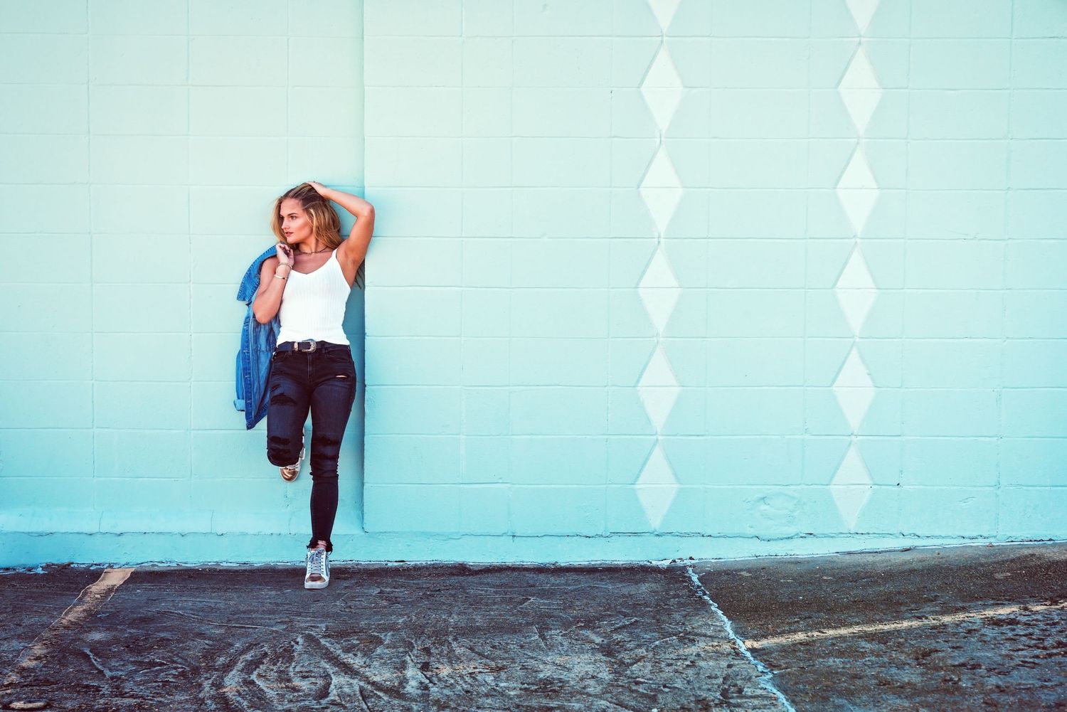 Young blonde woman leaning against a light blue wall outdoors holding a denim jacket casually.