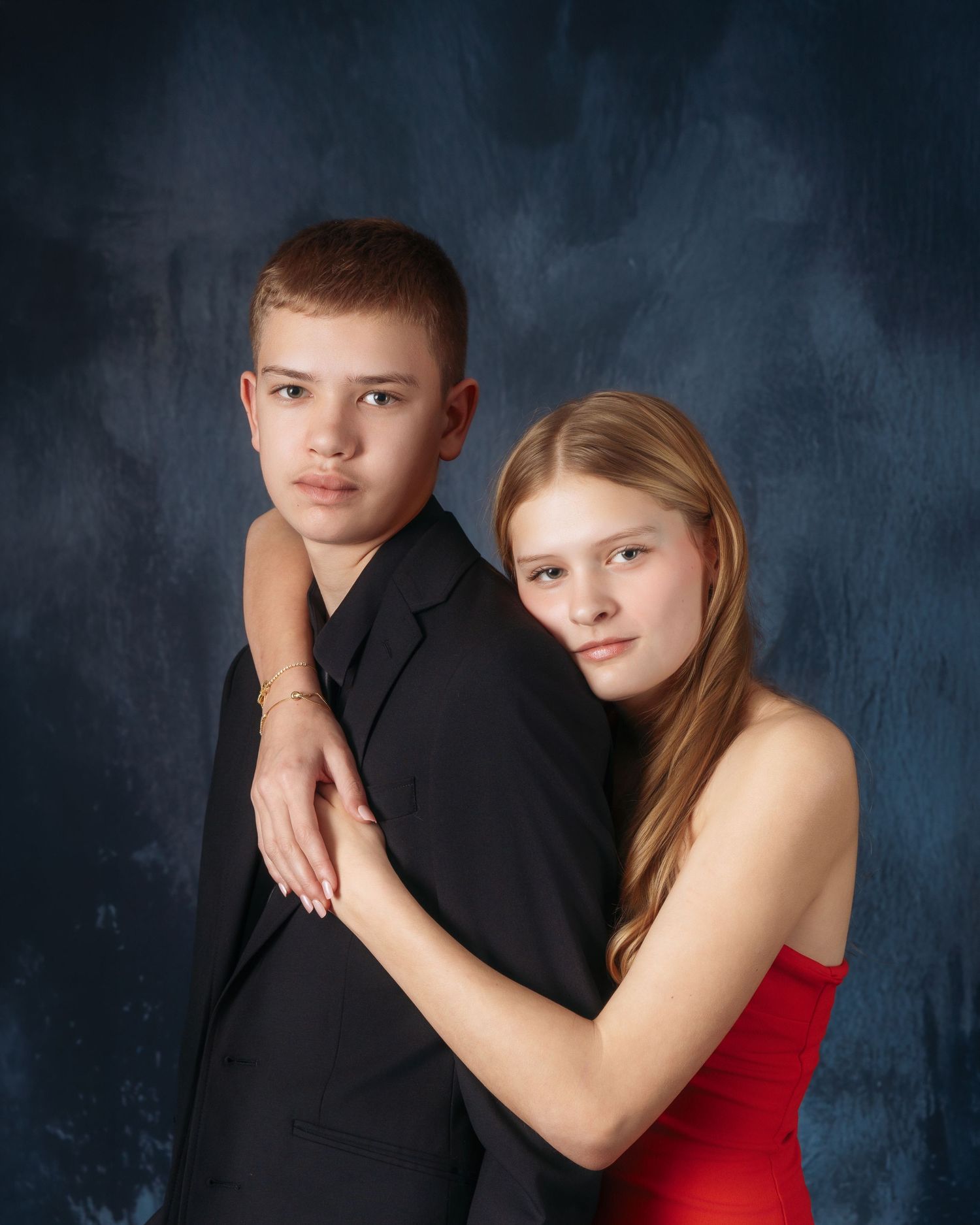 Two teenagers posing for a formal portrait, boy in black suit and girl in red dress, against blue background.