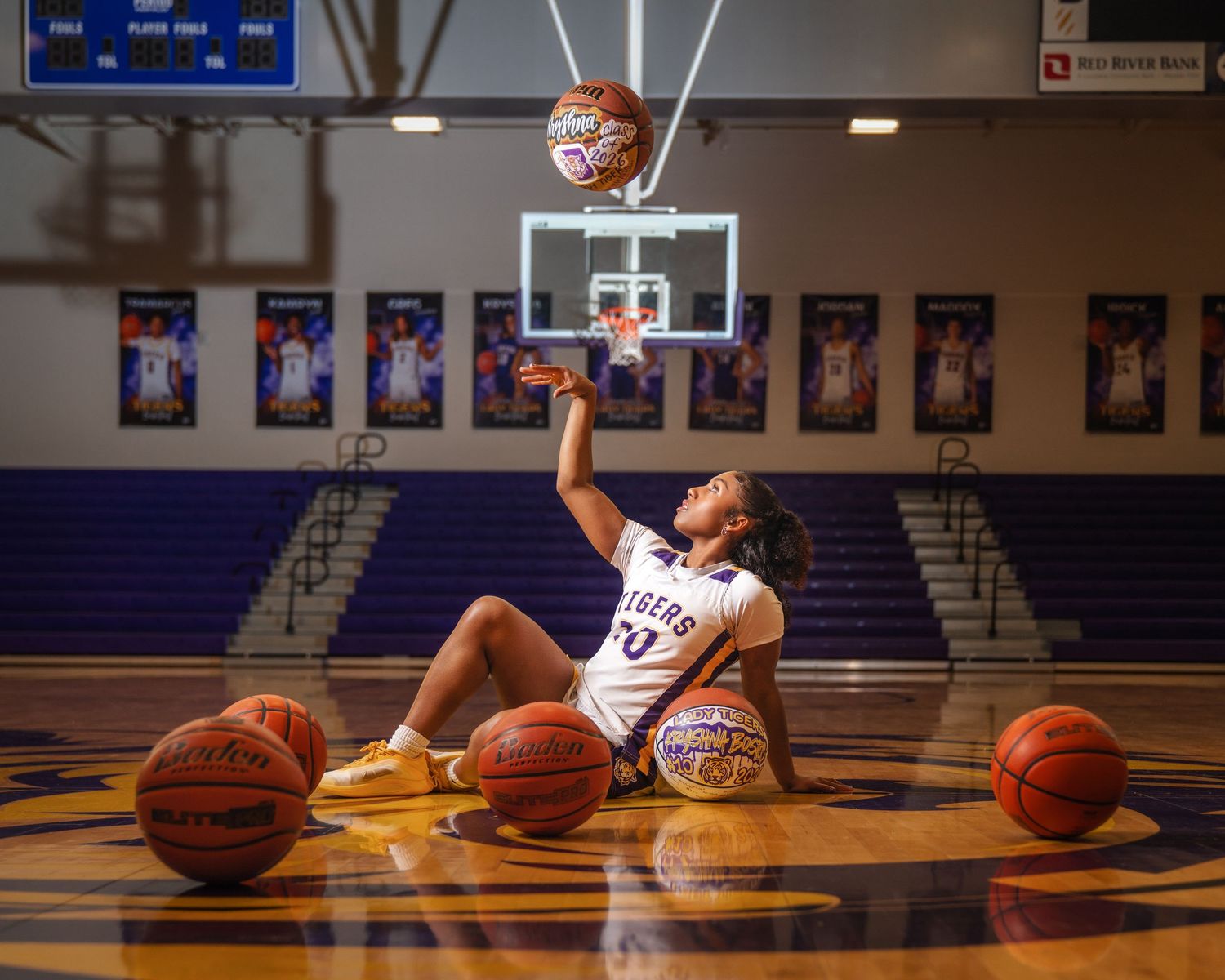 Female basketball player sits on court surrounded by balls, tossing one up toward hoop in purple gym.