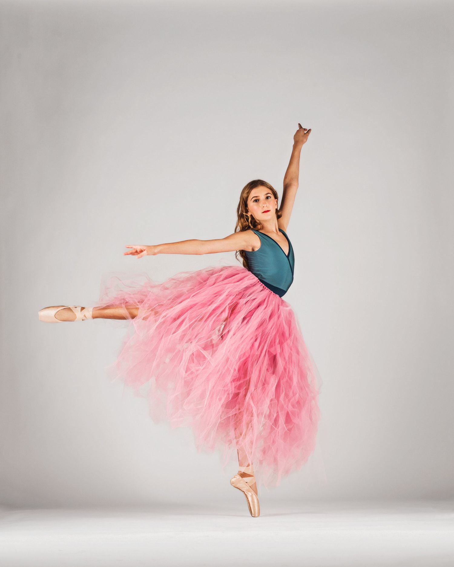 Ballerina in pink tutu and teal leotard performs an arabesque on pointe shoes against a white background.