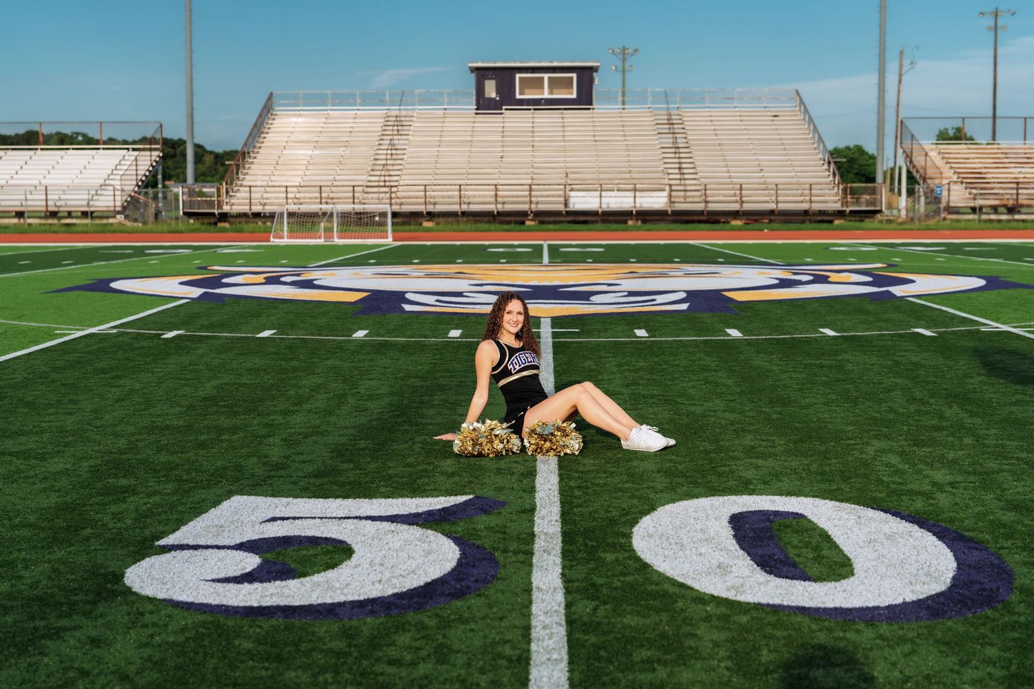 Cheerleader poses on the 50-yard line of a football field with pom-poms, bleachers visible in background.