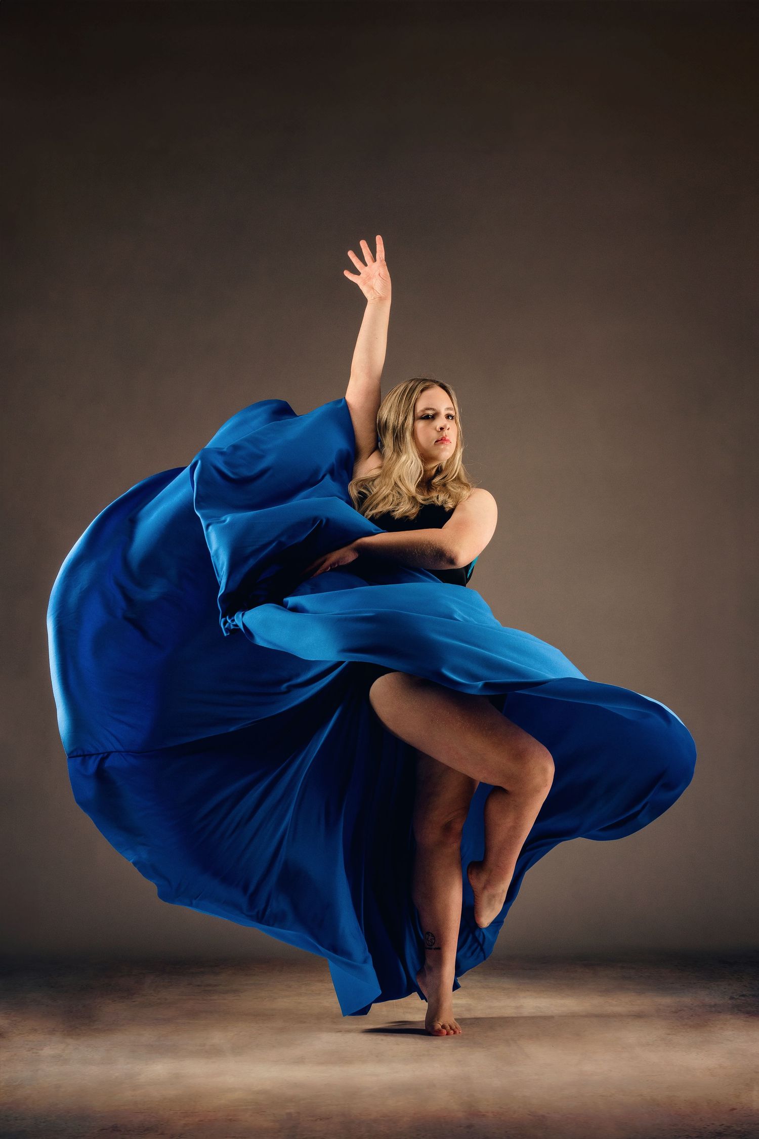 Blonde dancer in flowing blue gown mid-performance, arm raised gracefully against a dark studio background.