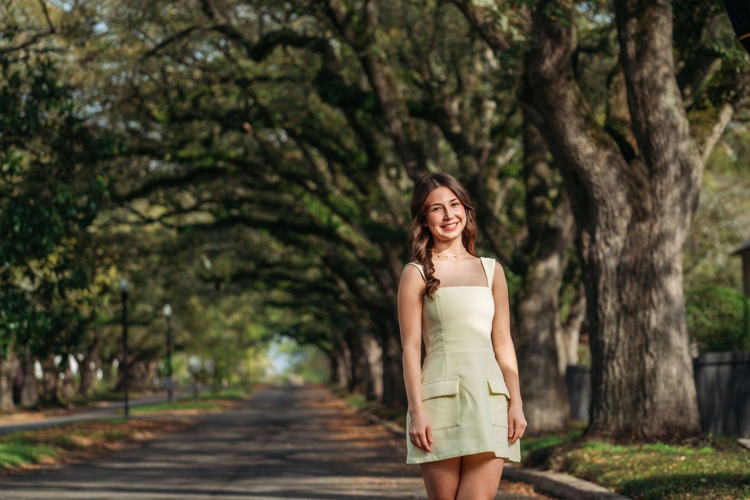 Young woman in a light green mini dress smiling on a tree-lined road in a sunny outdoor setting.