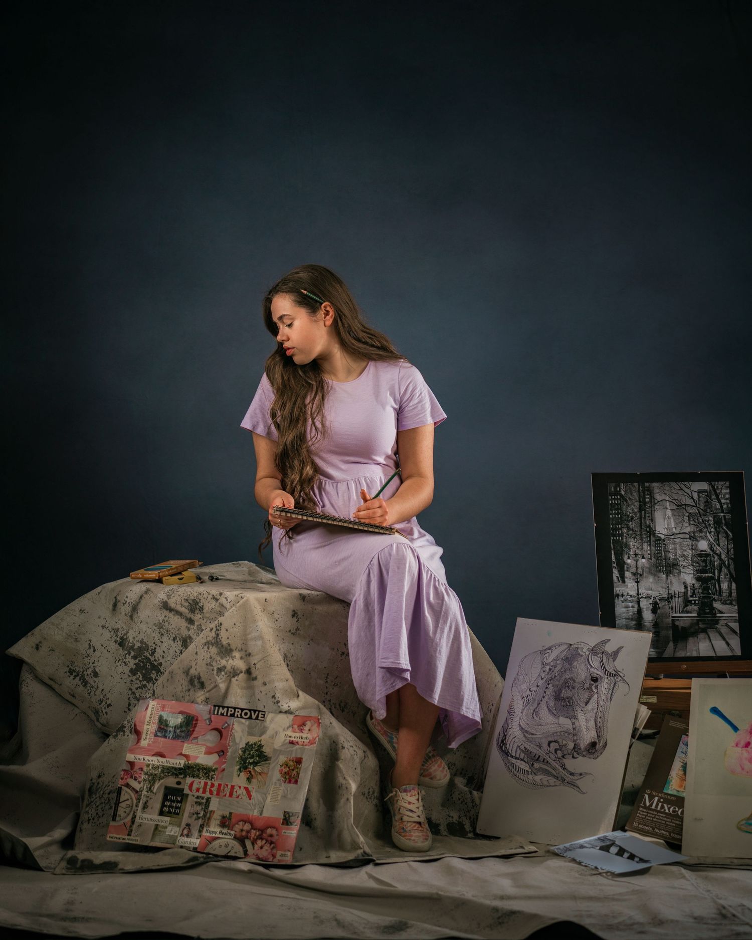 Young woman in lavender dress sketching artwork, surrounded by drawings and paintings on a dark studio backdrop.