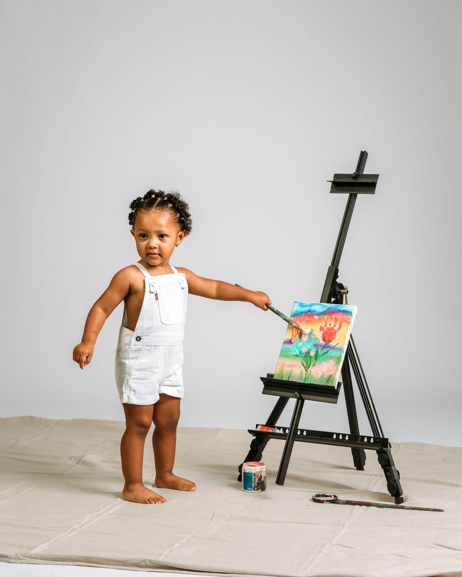 Toddler in white overalls painting on an easel with colorful artwork in a studio setting.