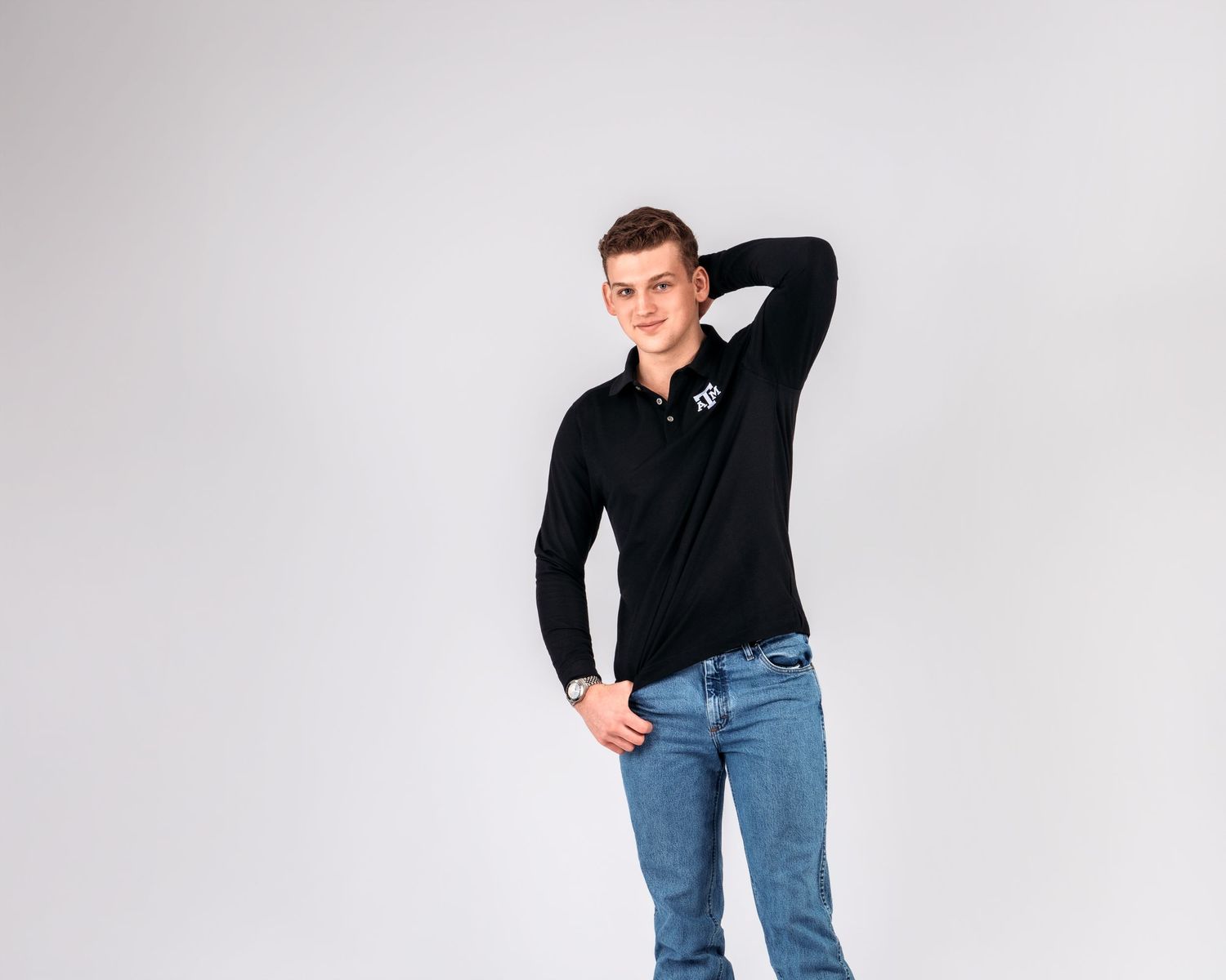 Young man in black long-sleeve polo and jeans posing confidently against a light gray background.