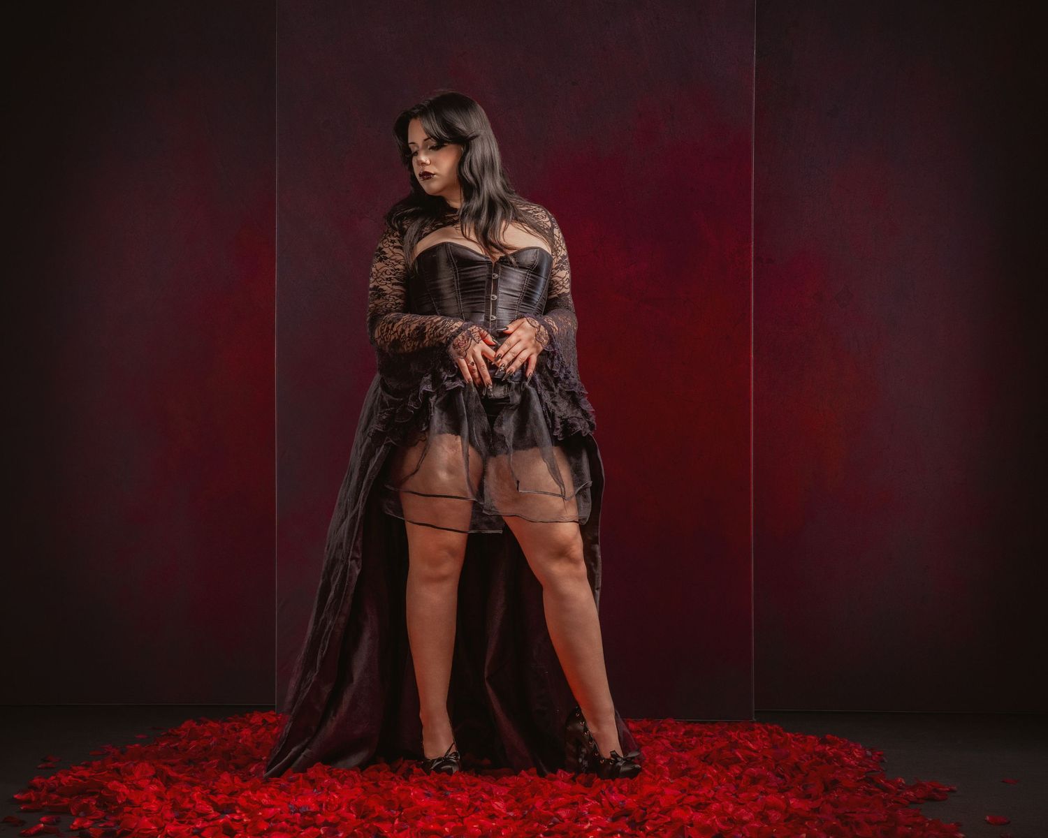 Woman in black corset and sheer robe poses dramatically on red rose petals against dark crimson backdrop.