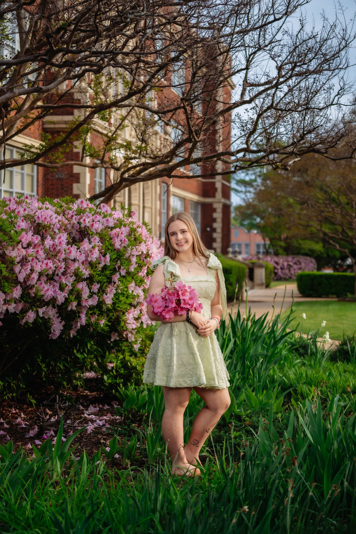 Young blonde woman in a green dress holding pink flowers near blooming azaleas outside a brick building.