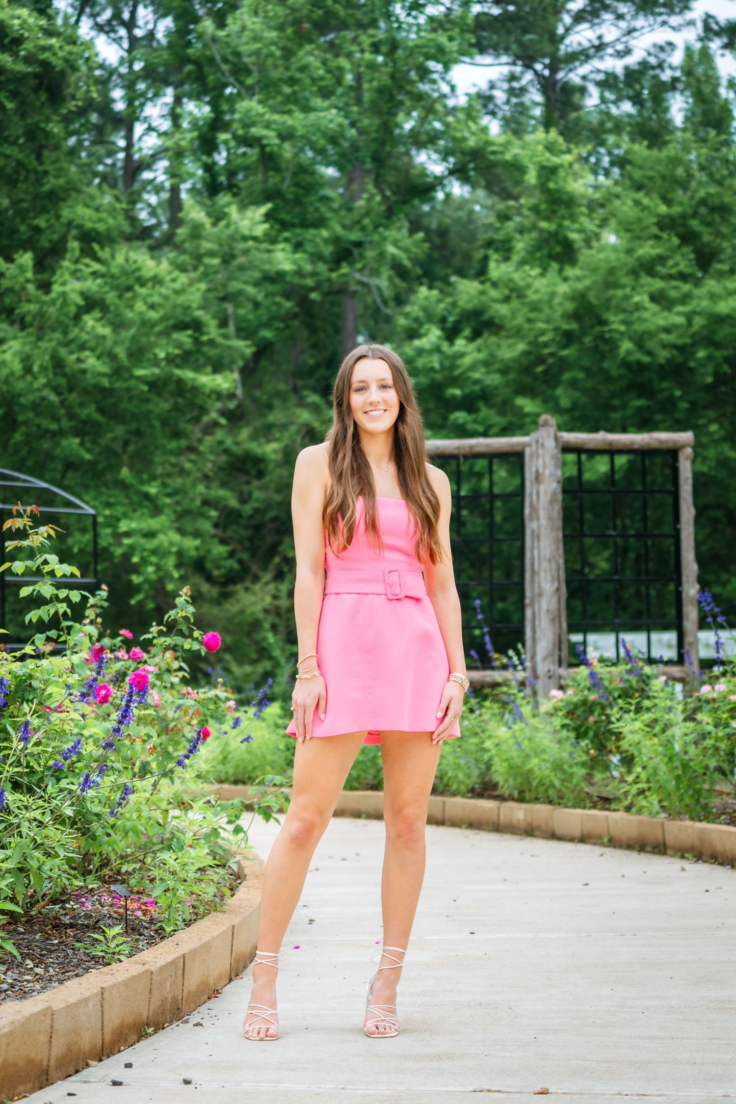 Young woman in pink mini dress posing in garden with colorful flowers and green trees.