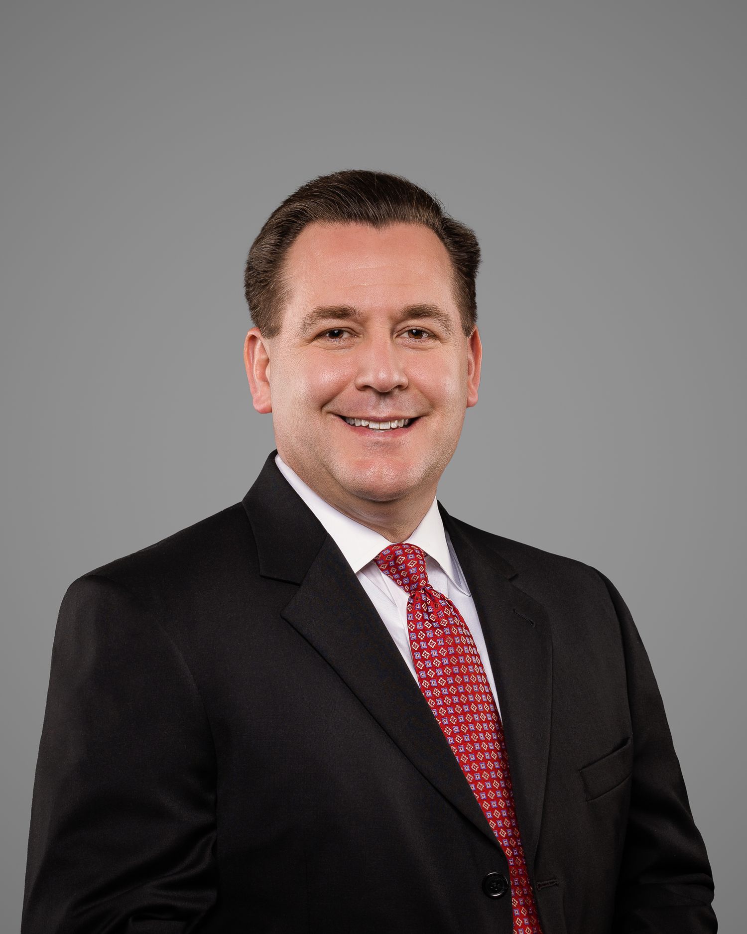 Professional business headshot of man in black suit and red tie smiling confidently.