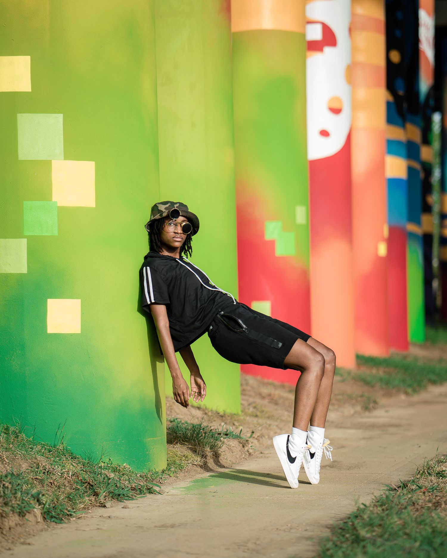 Young dancer in black outfit leans against colorful painted pillars in urban outdoor setting.