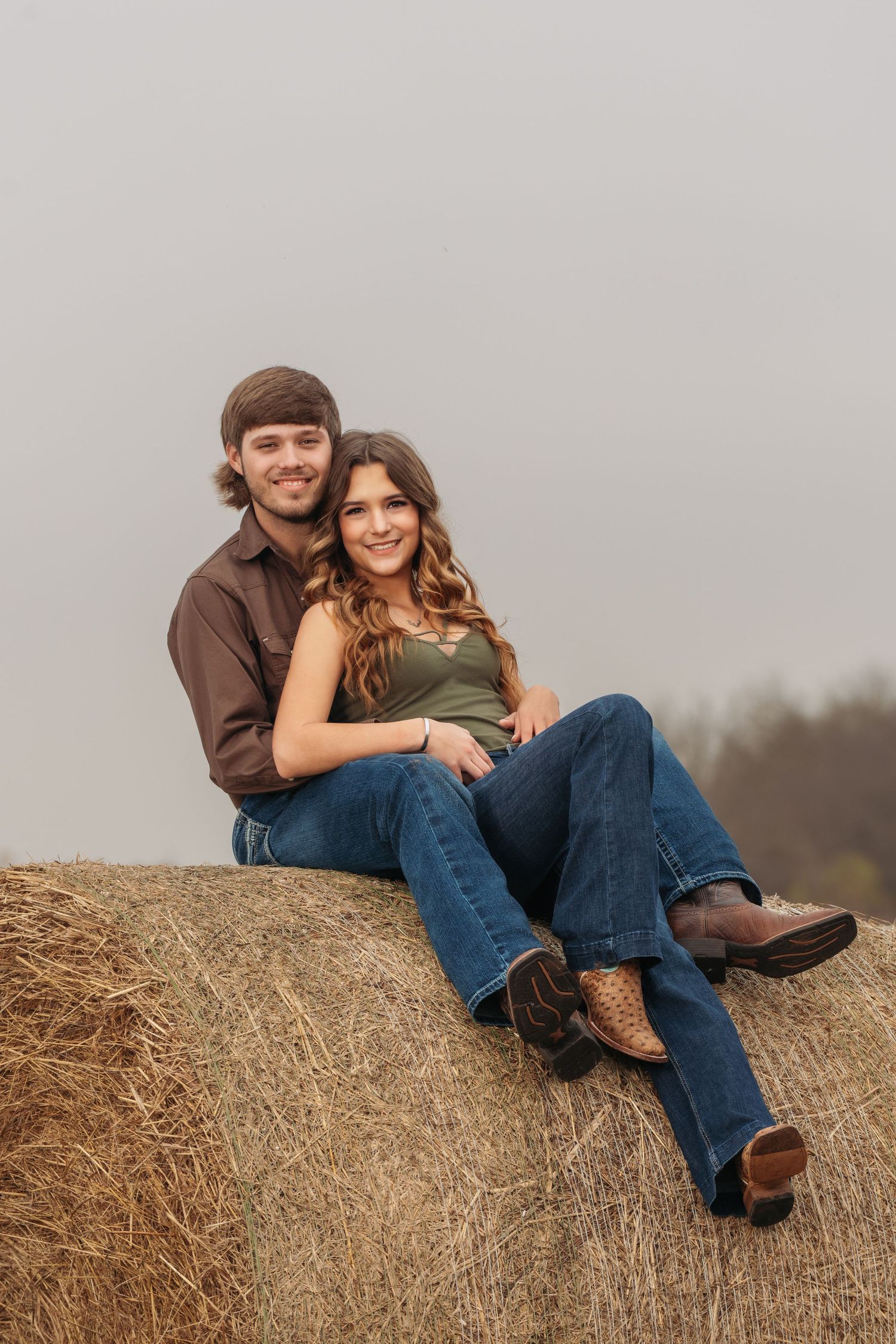 Smiling couple sitting together on a large hay bale in a misty rural field.