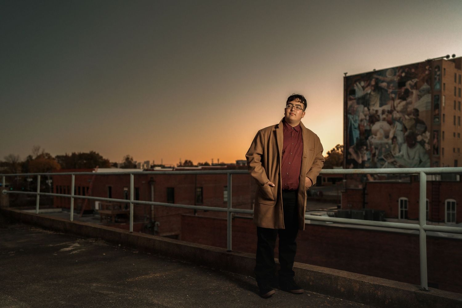 Man in trench coat and beanie stands on rooftop at sunset with urban mural and brick buildings behind him.