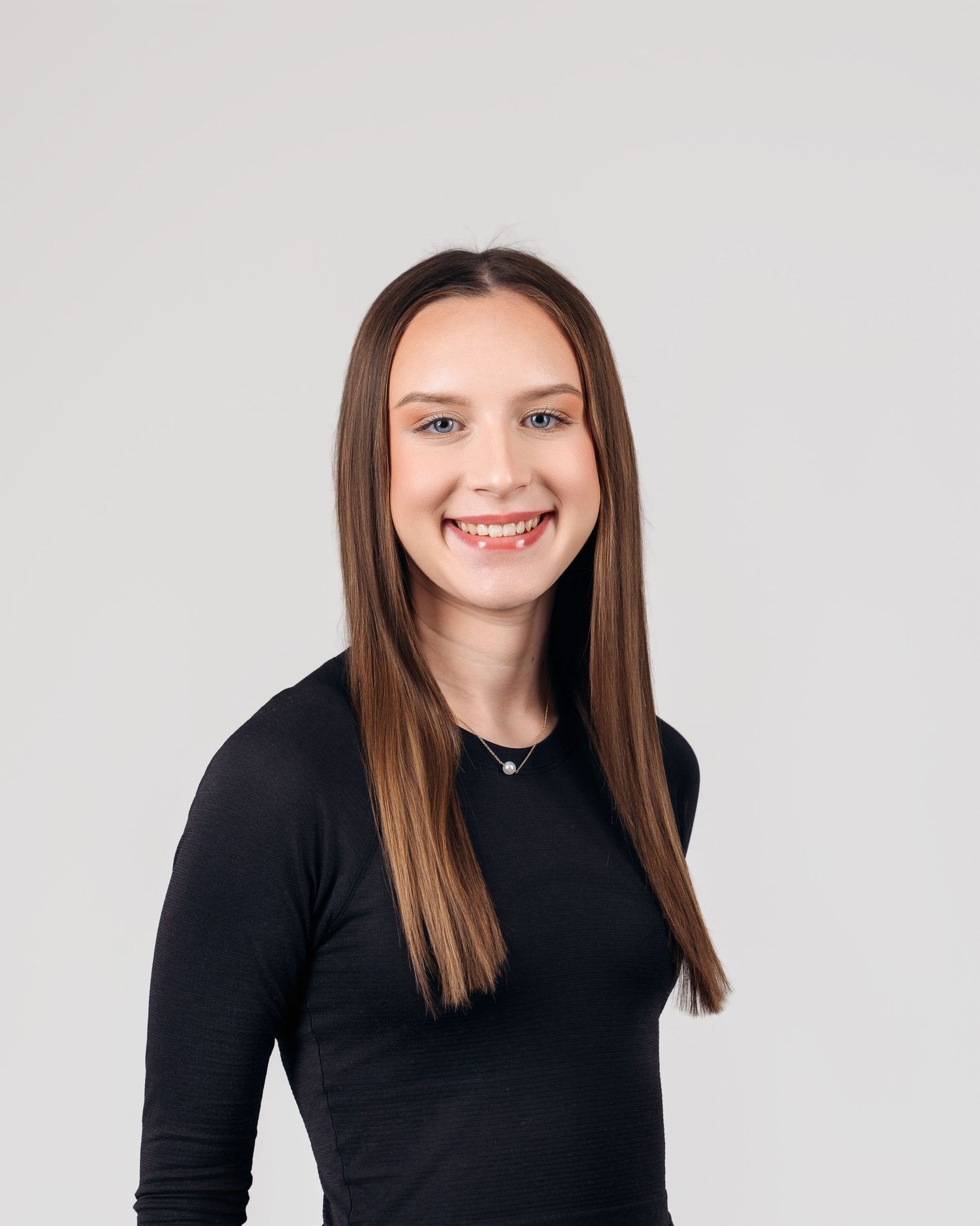Young woman with long brown hair smiling, wearing a black long-sleeve top and small necklace against a gray background.