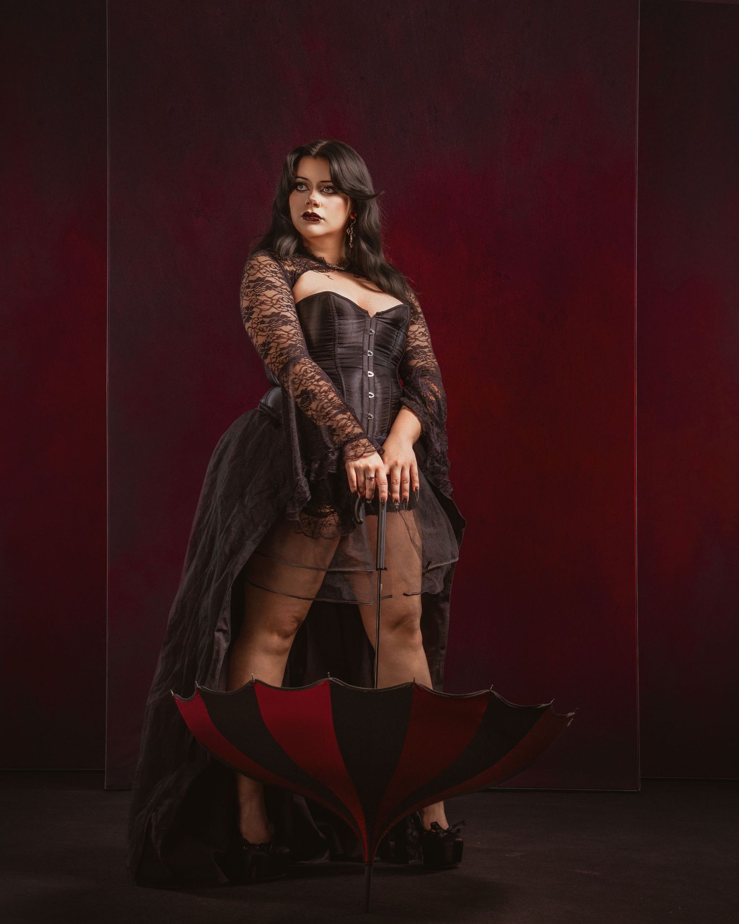 Woman in gothic corset dress with lace sleeves poses dramatically with red and black umbrella against dark crimson backdrop.