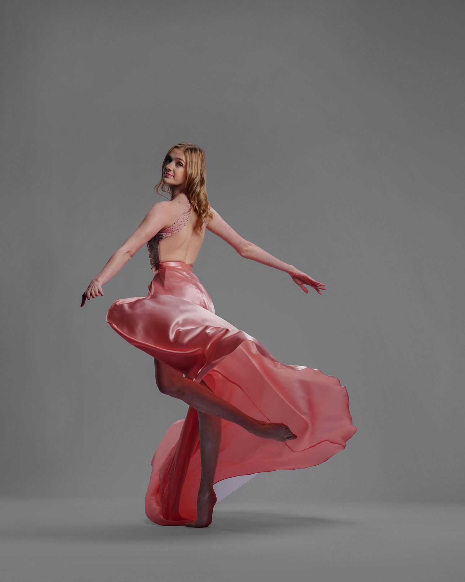 A woman in a flowing red satin backless gown twirls gracefully against a gray studio background.